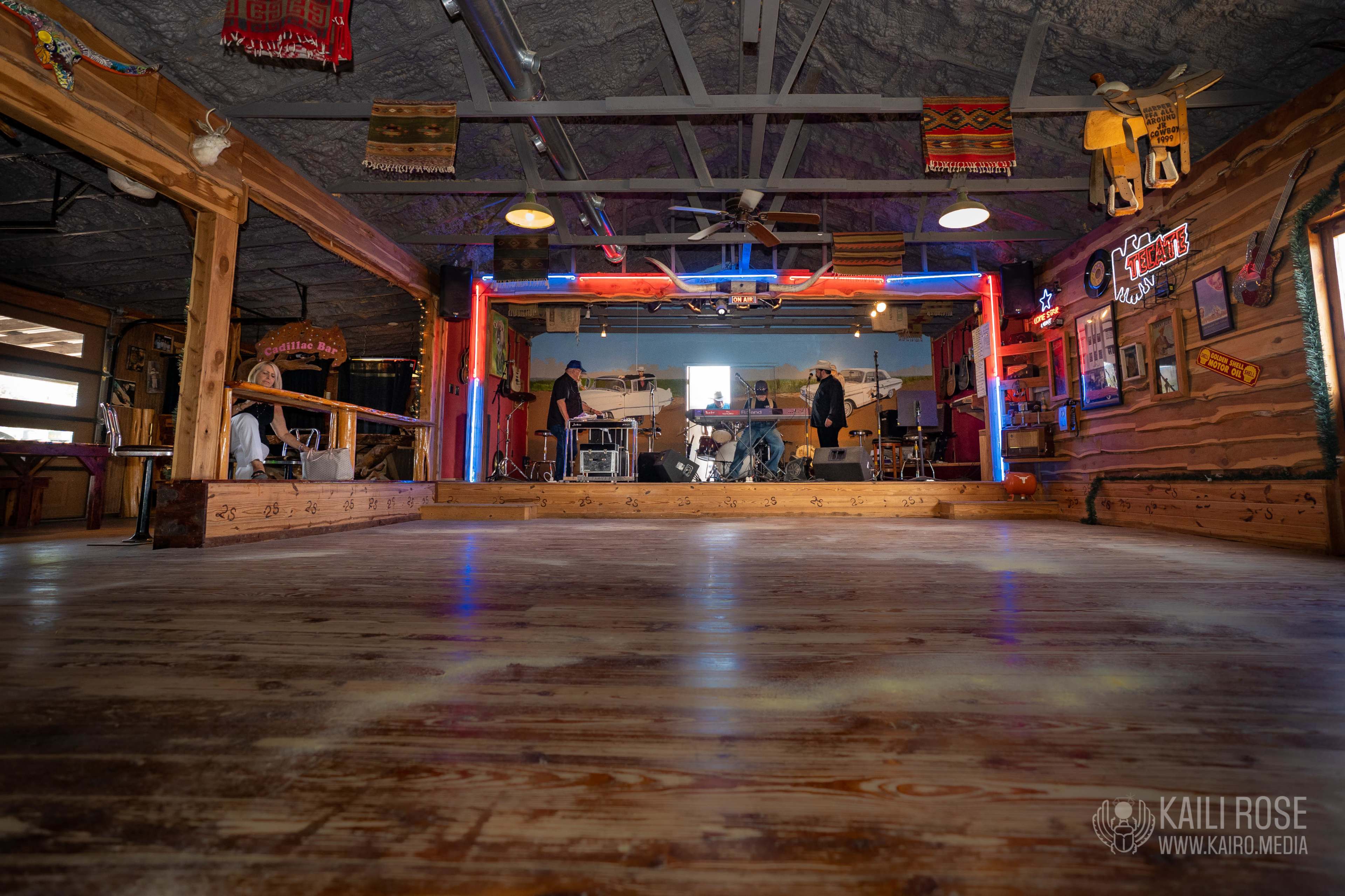 A wooden dance hall with a raised stage, colorful neon lights, and a band preparing to perform in the background.