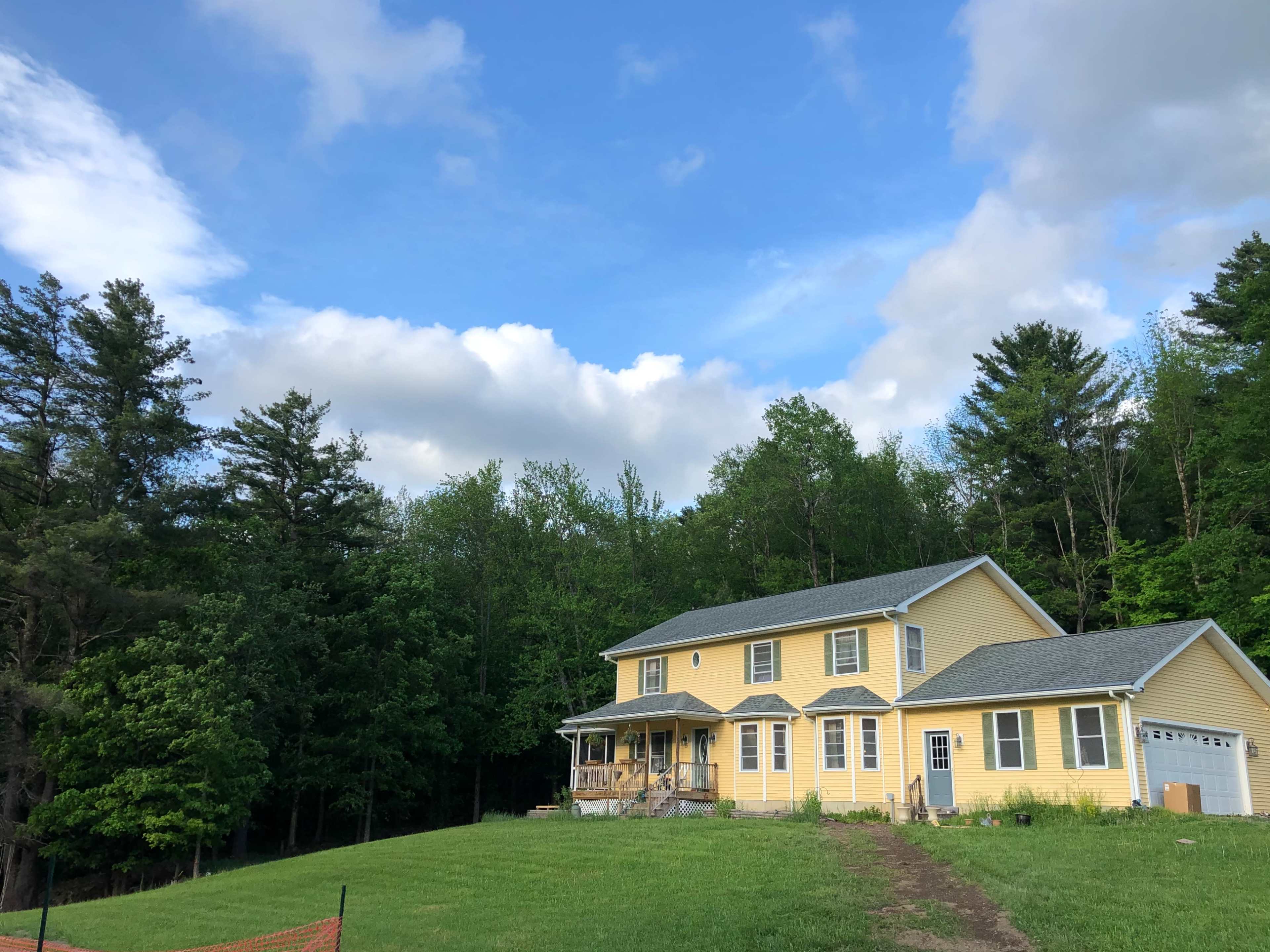 A two-story yellow house is situated near a wooded area under a partly cloudy sky.