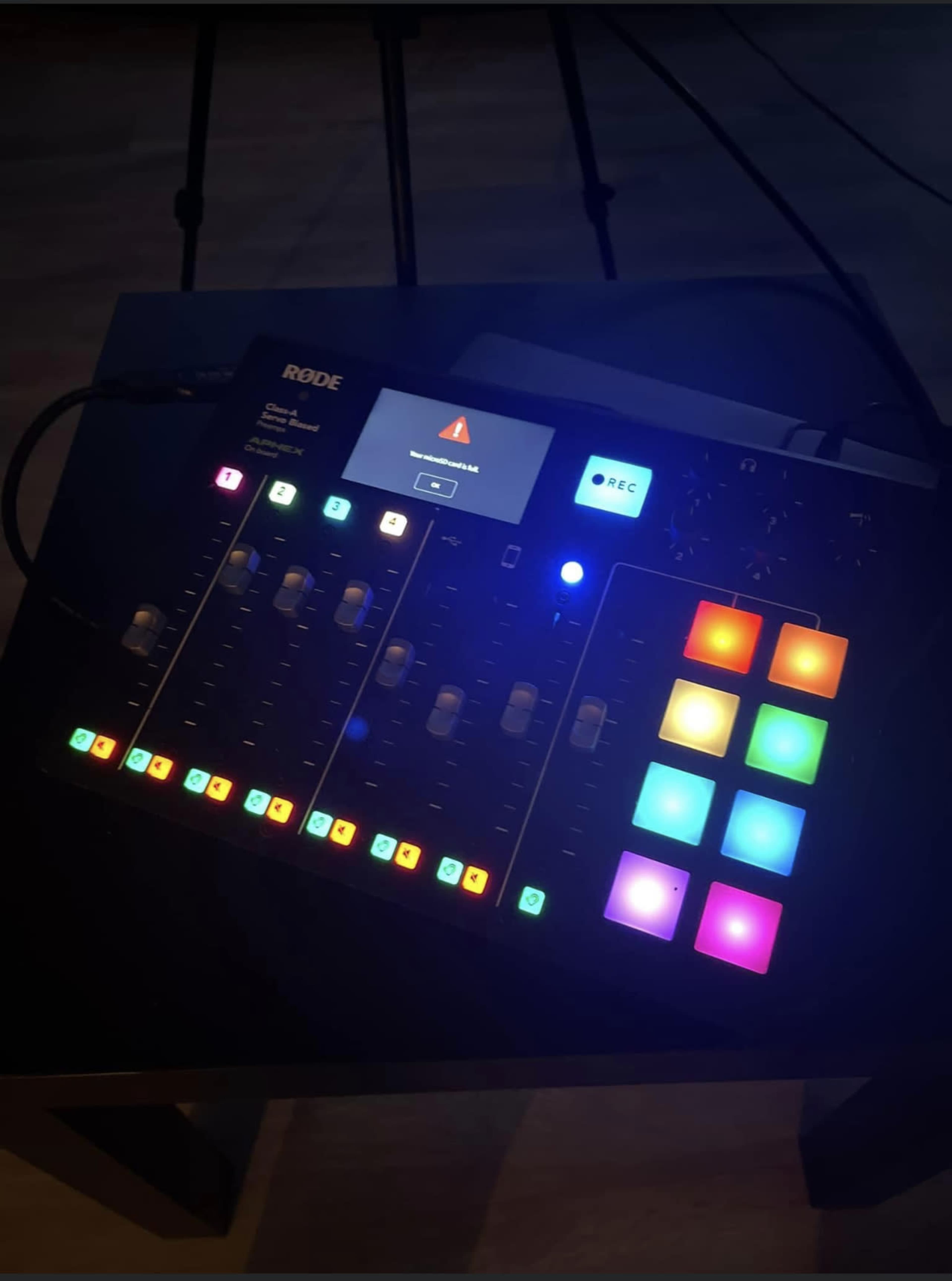 The image shows a Rode audio mixer with various sliders, buttons, and a display screen, illuminated with multicolored lights.
