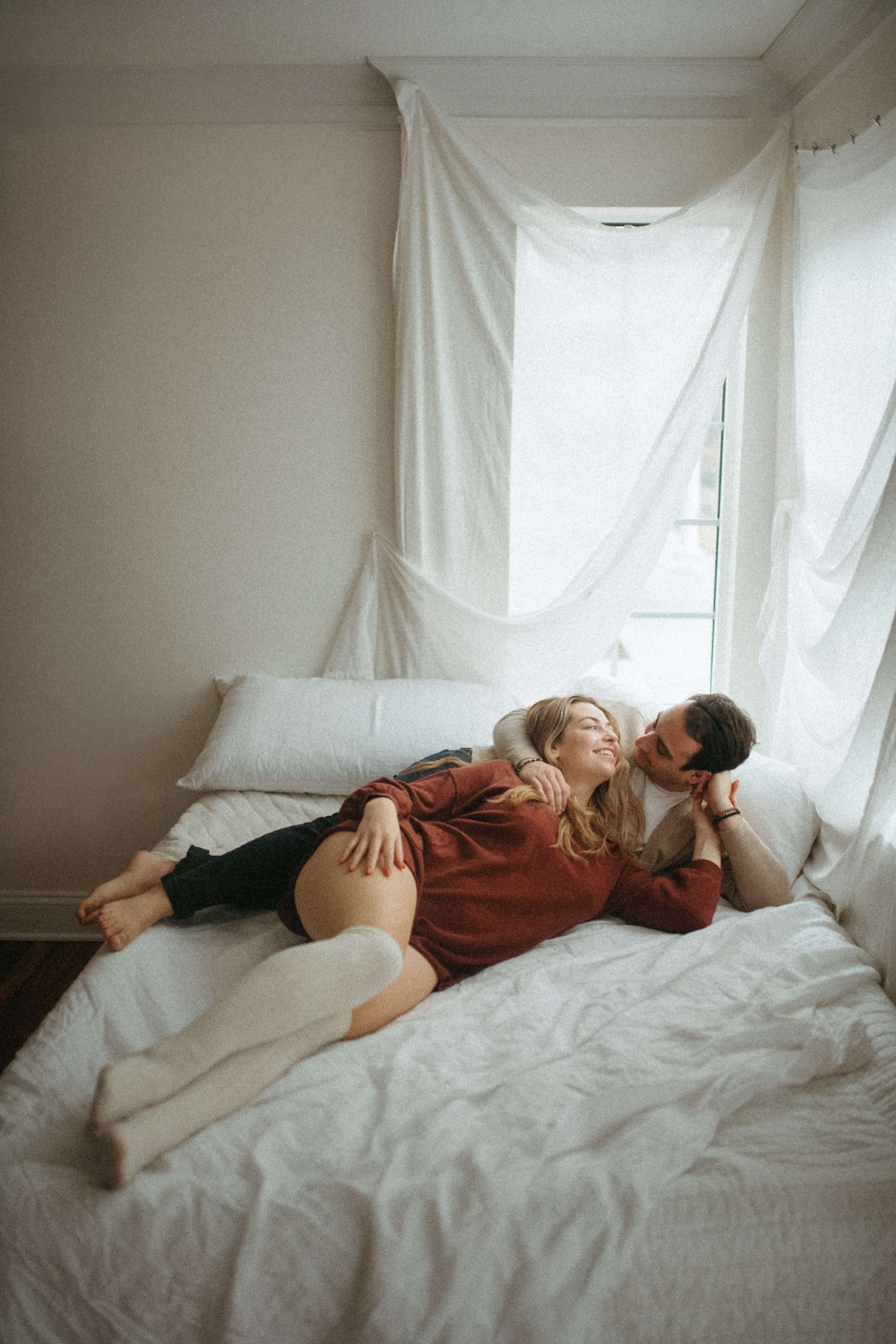 A couple lies together on a bed, surrounded by light-colored blankets and draped fabric, engaging in a tender moment.