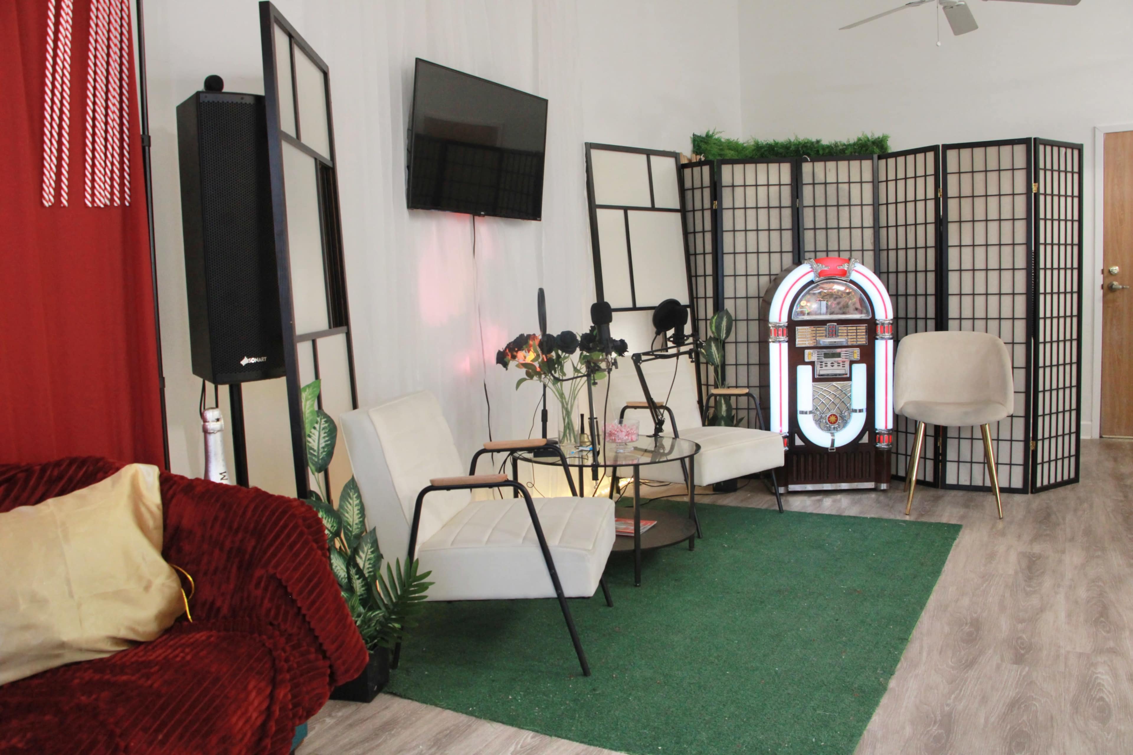 The image shows a cozy lounge area featuring white chairs, a small glass table, a jukebox, and decorative plants, set against a backdrop of red curtains and a wooden partition.
