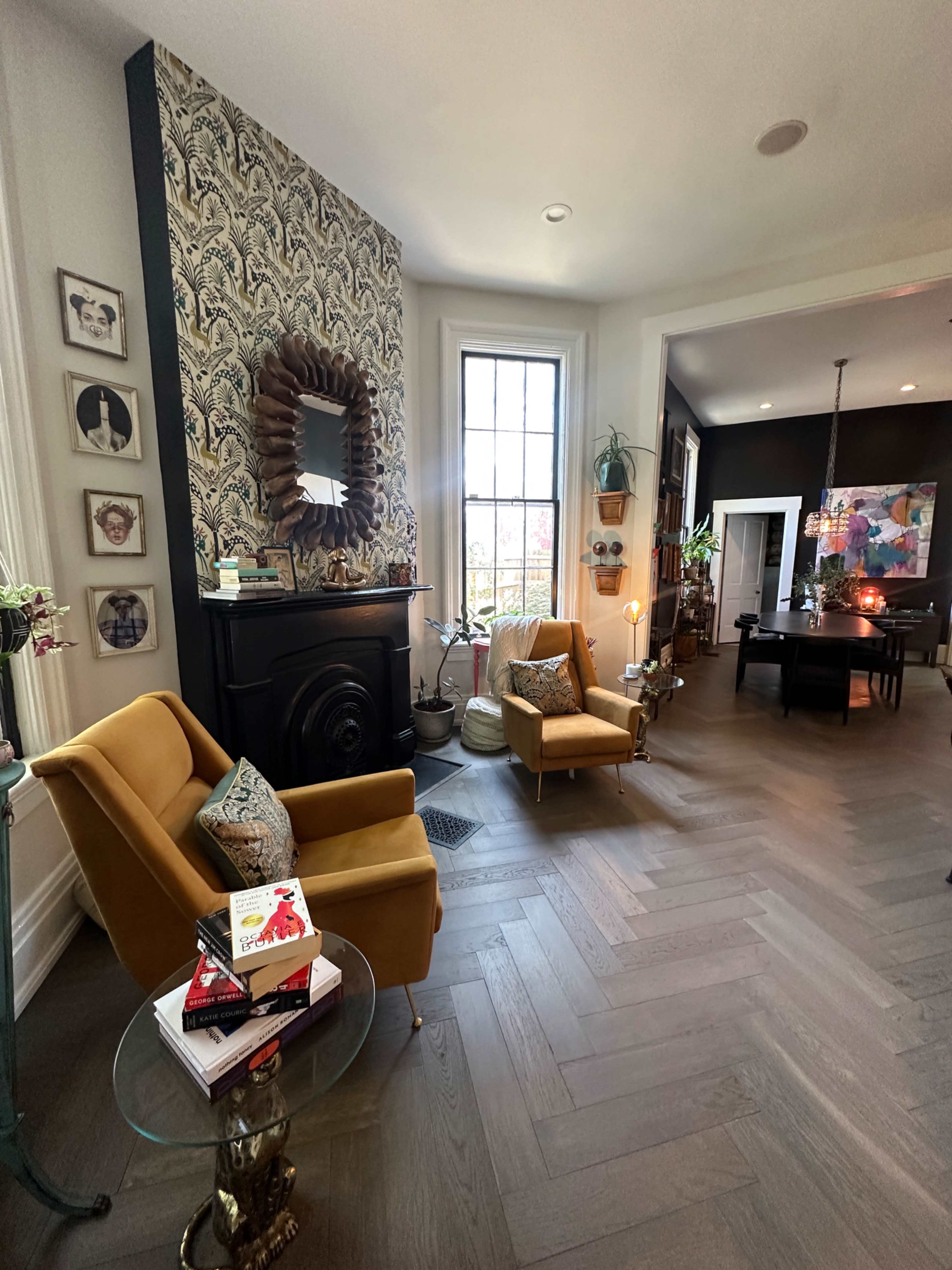 The image shows a stylish living room featuring two chairs, a round side table with books, a decorative mirror, and a dining area visible in the background.