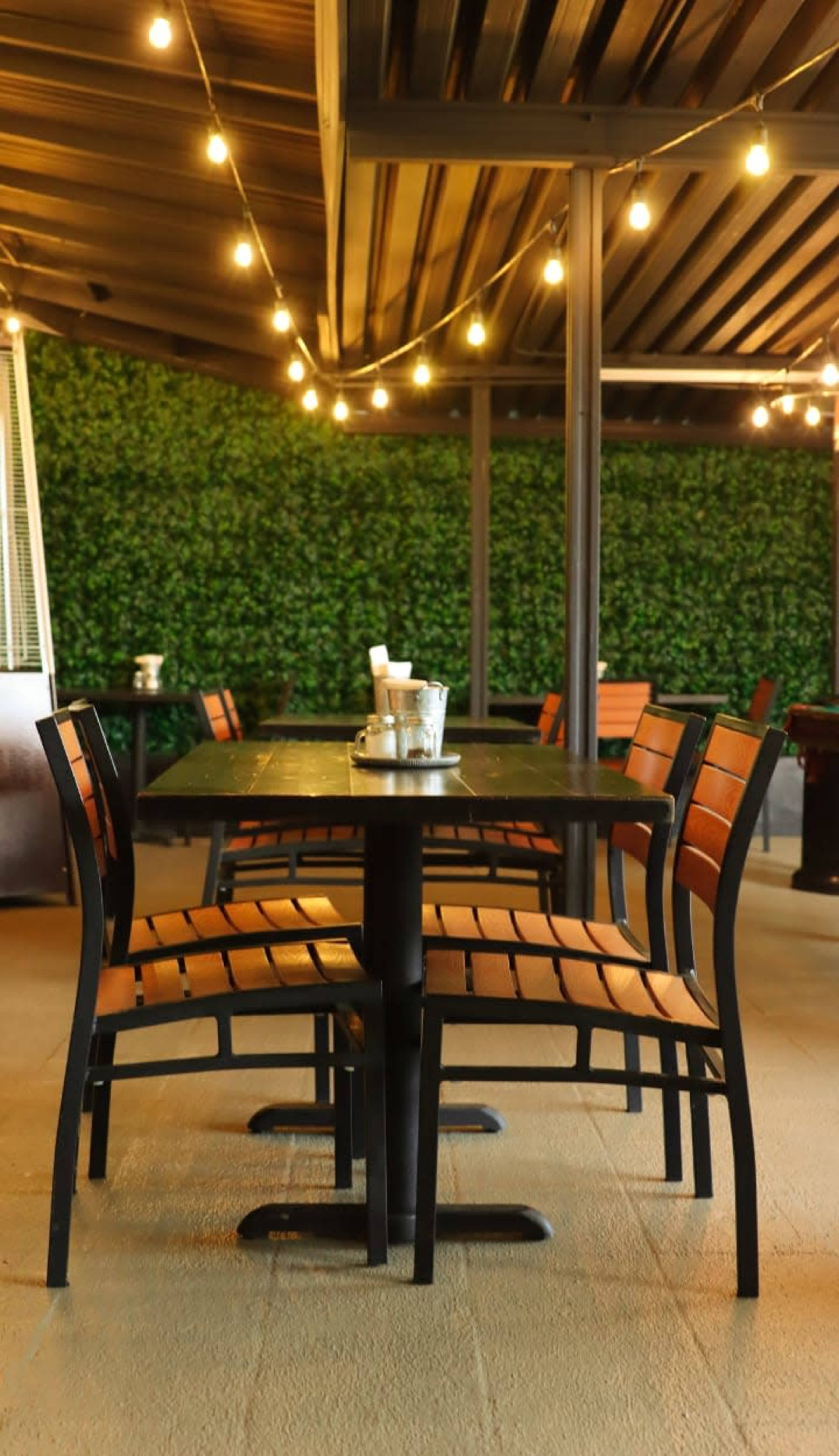 A table with four chairs is set in a well-lit outdoor dining area surrounded by green foliage and string lights.