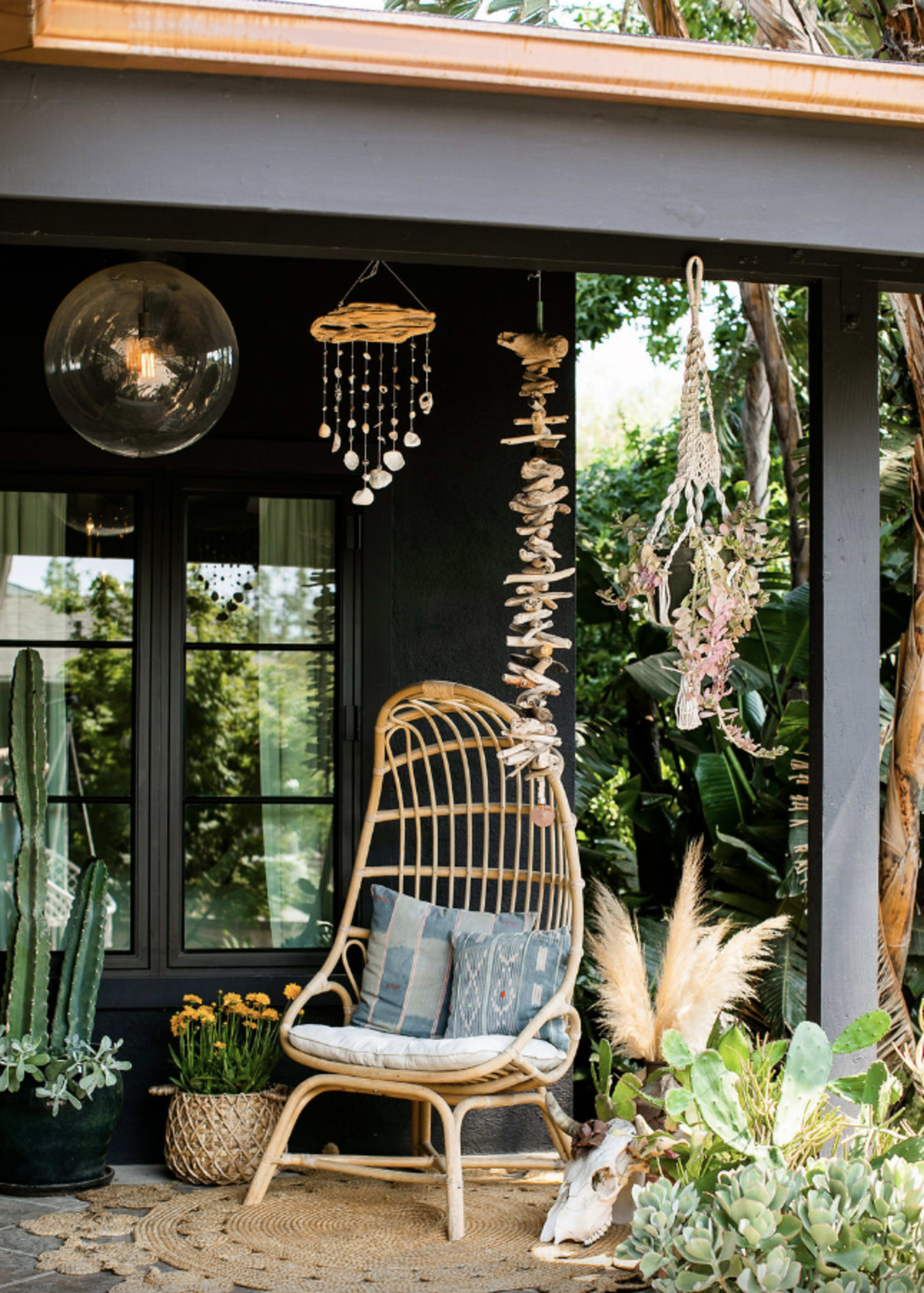 A rattan chair is placed on a porch surrounded by various plants, with hanging decorations and a round light fixture above.