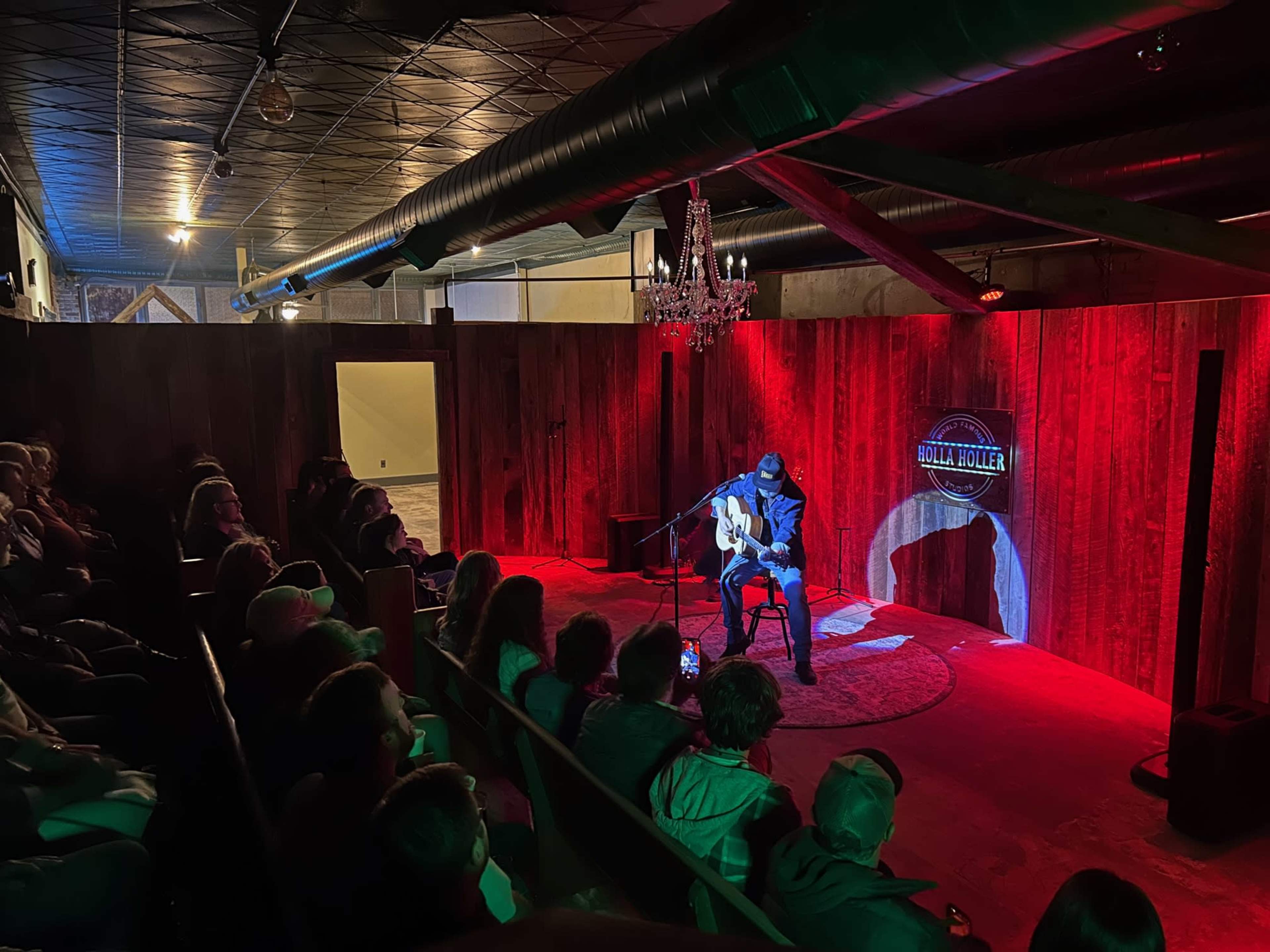 A performer plays guitar on stage in a dimly lit venue while an audience watches from the seats.