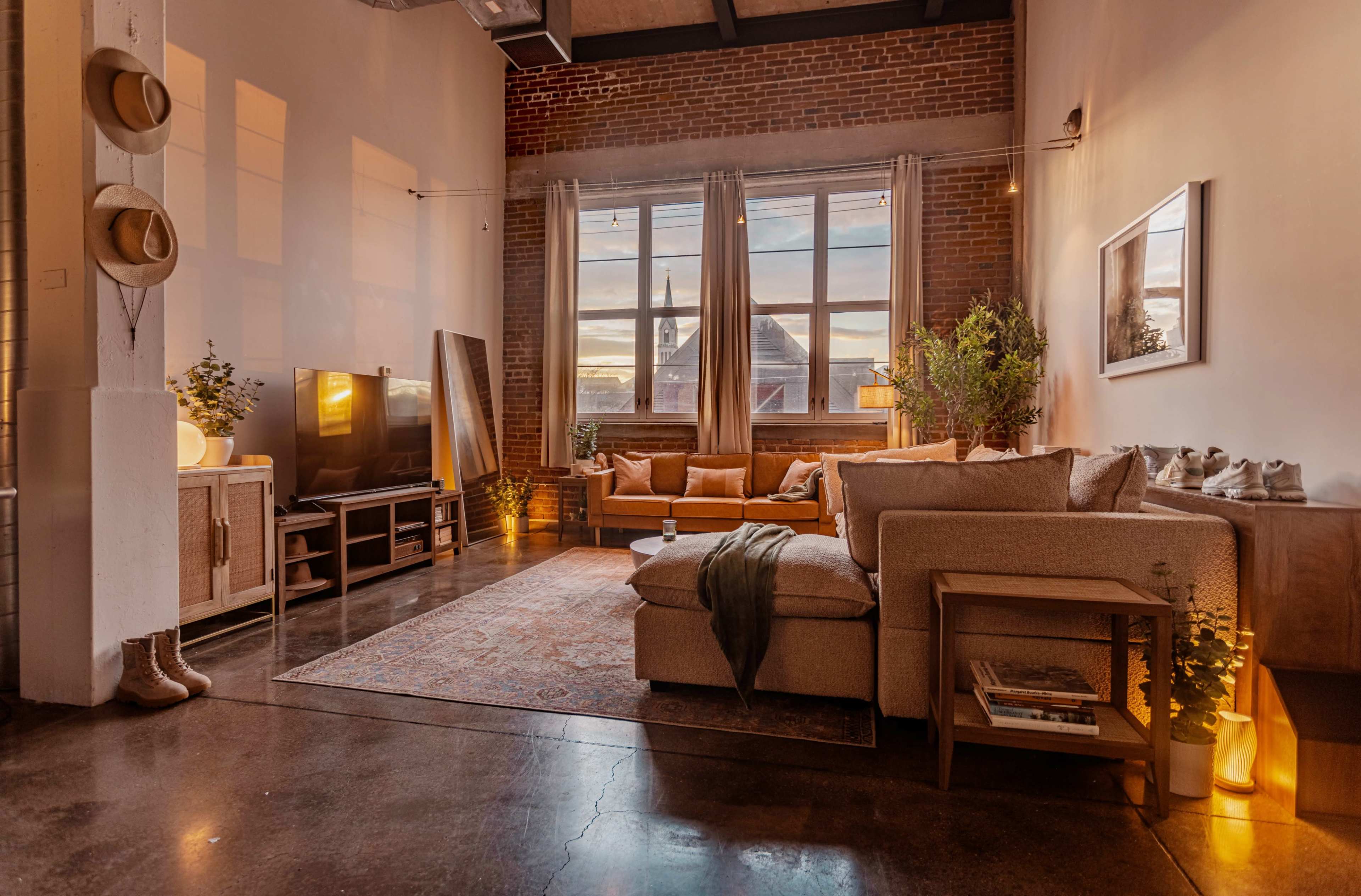 The image shows a cozy living room with large windows, exposed brick walls, and a mix of modern furniture, including a sofa and a wooden coffee table, illuminated by warm ambient lighting.