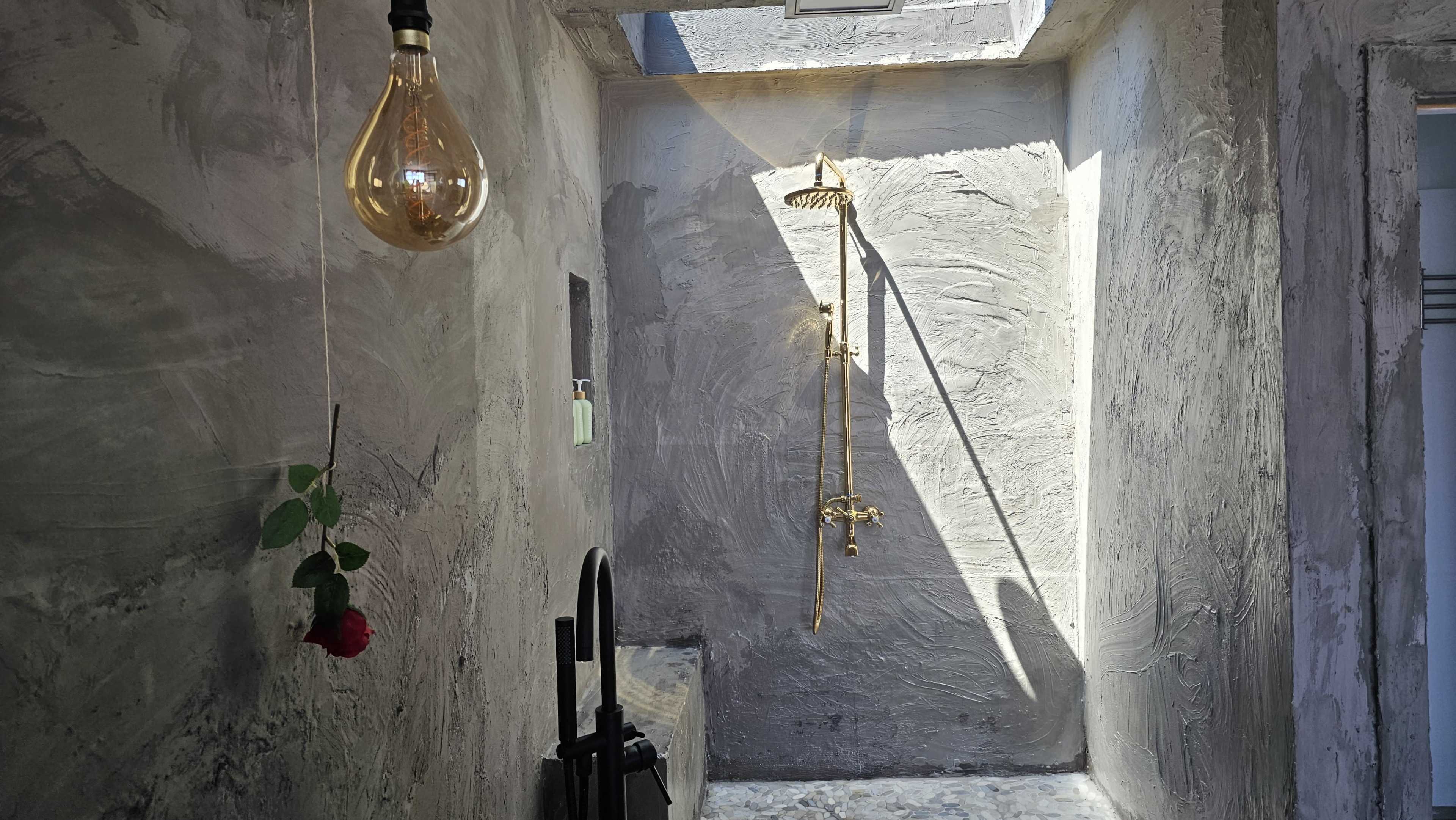 The image shows a modern shower area with textured gray walls, a gold shower fixture, and a hanging light bulb above a single rose.