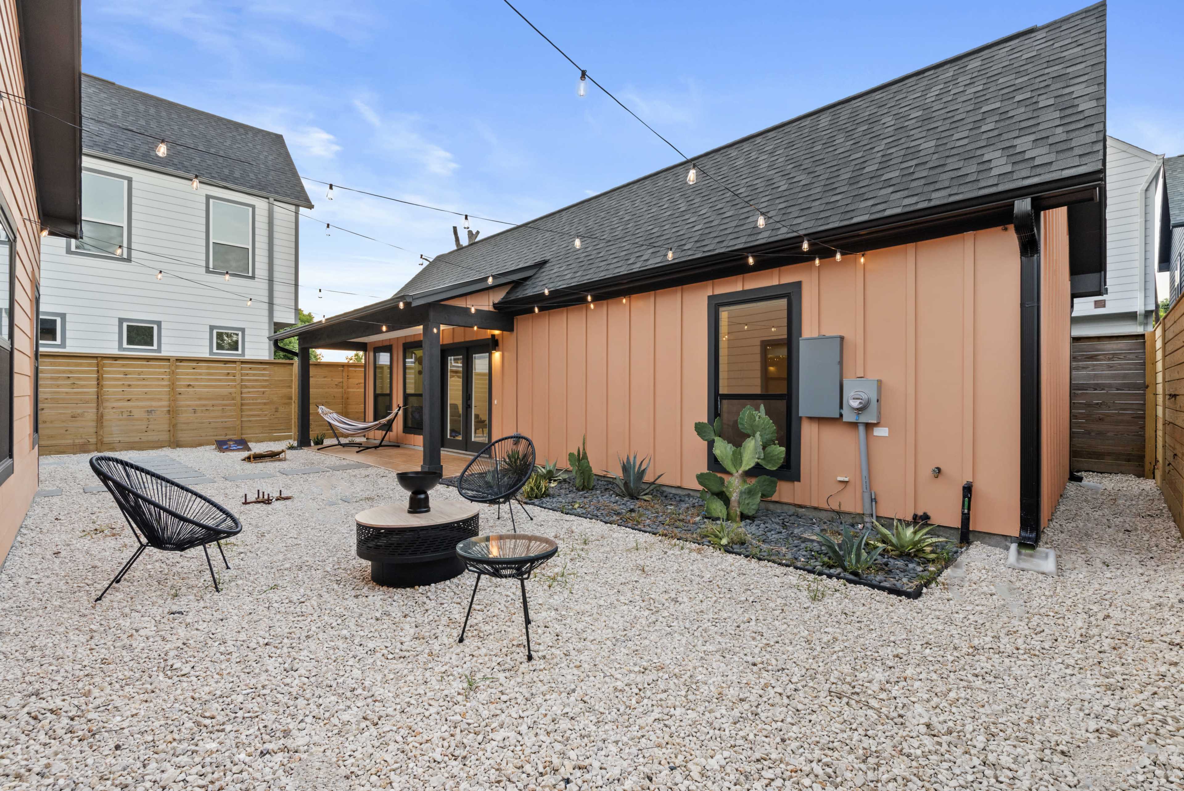 The image shows a gravel courtyard featuring a fire pit, modern chairs, and various plants, surrounded by buildings with contrasting exterior colors.
