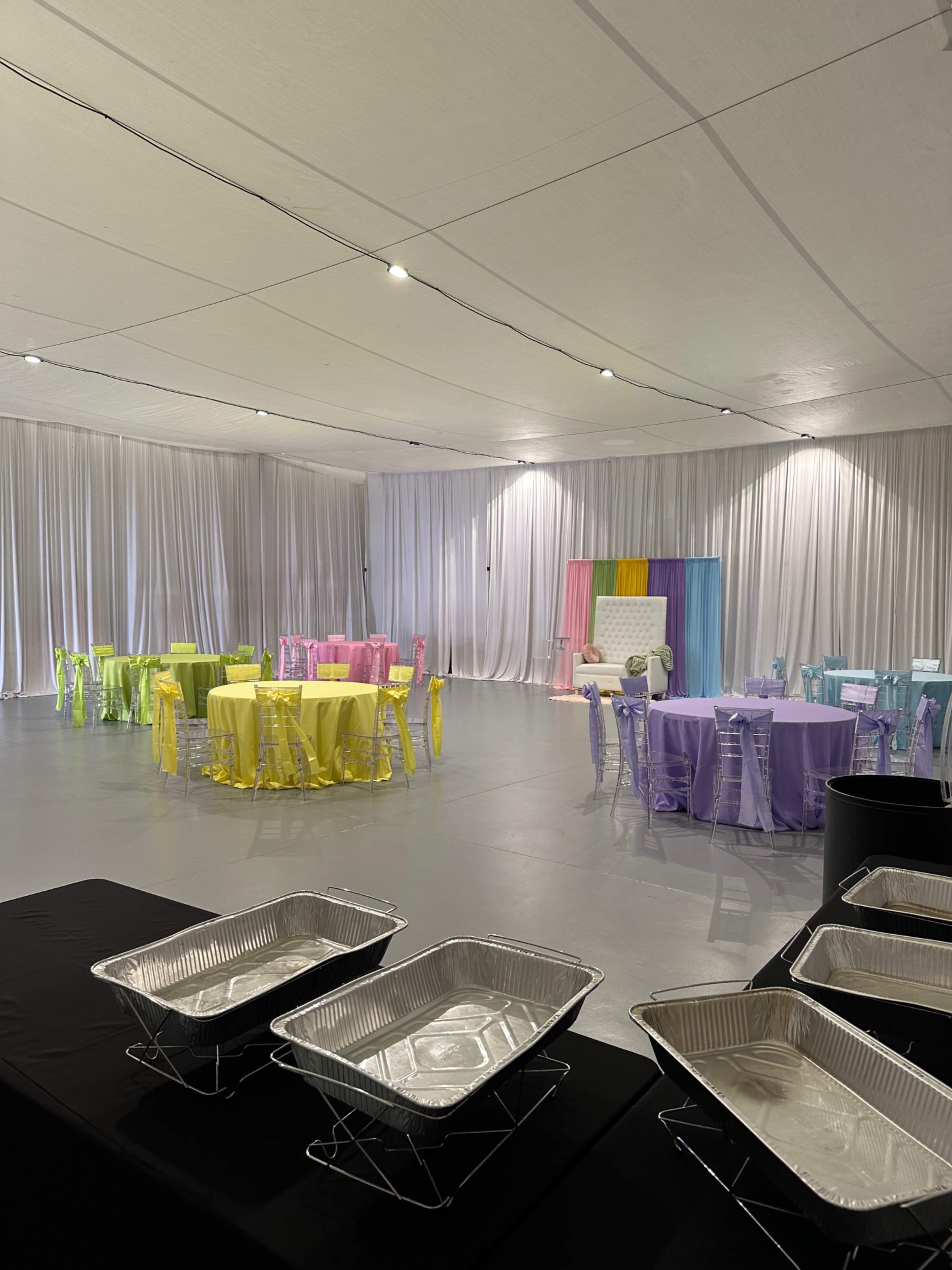 The image shows an event space set up with tables covered in pastel-colored tablecloths and some silver serving trays in the foreground.