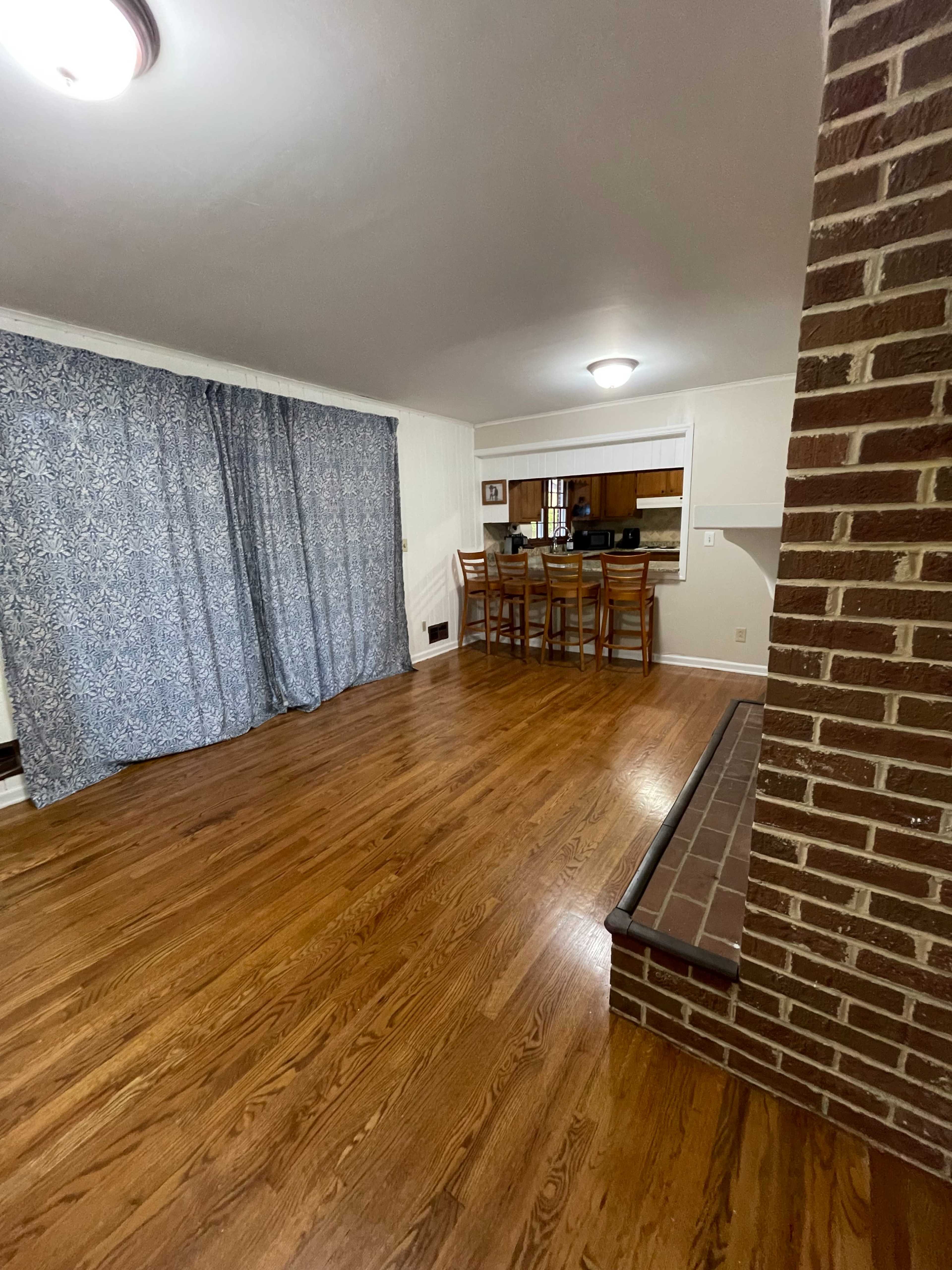 A spacious living area features hardwood floors, a curtain draped to one side, and a dining set visible in the background.