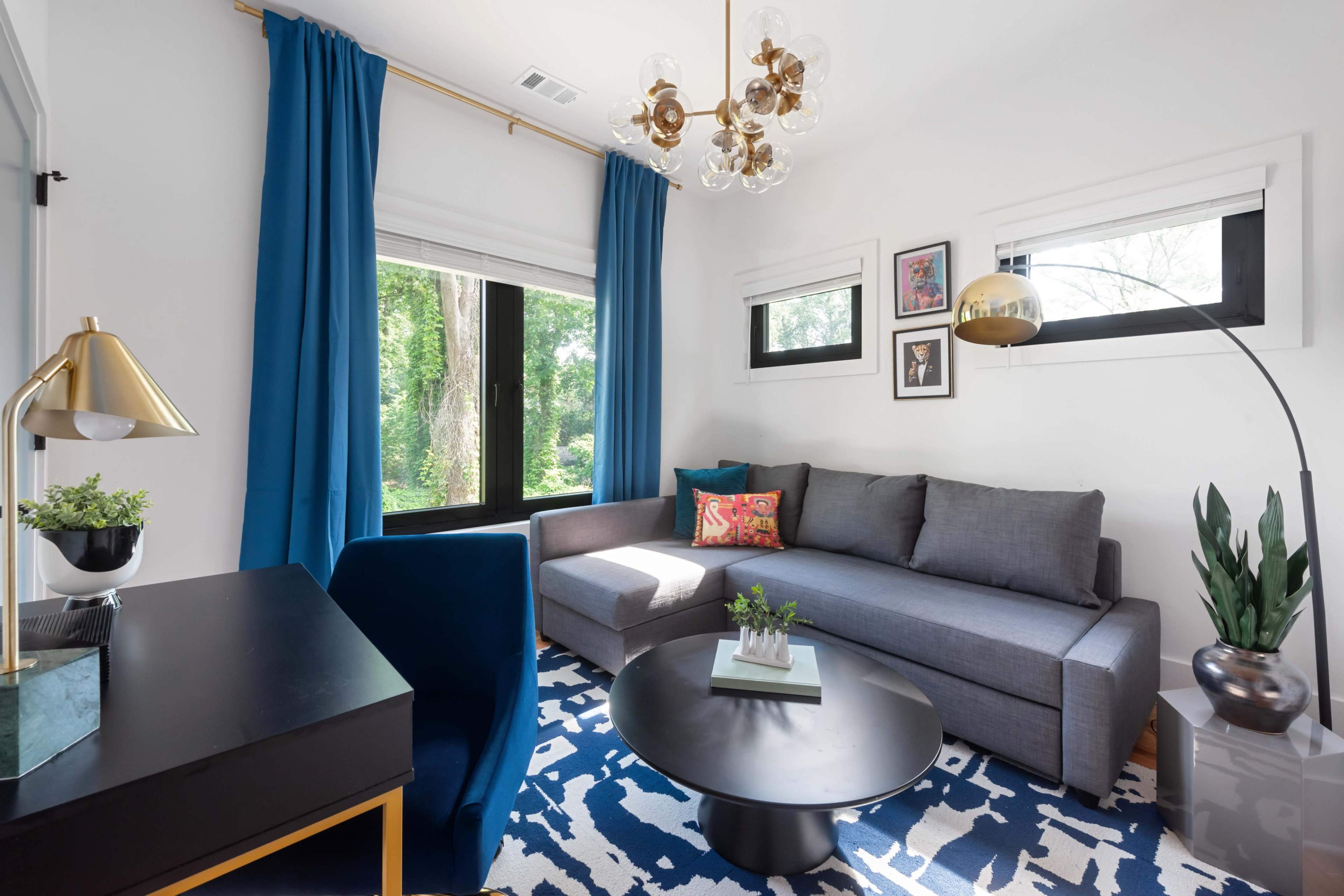 The image shows a modern living room featuring a gray sectional sofa, a round black coffee table, a blue and white patterned rug, and large windows with blue curtains.