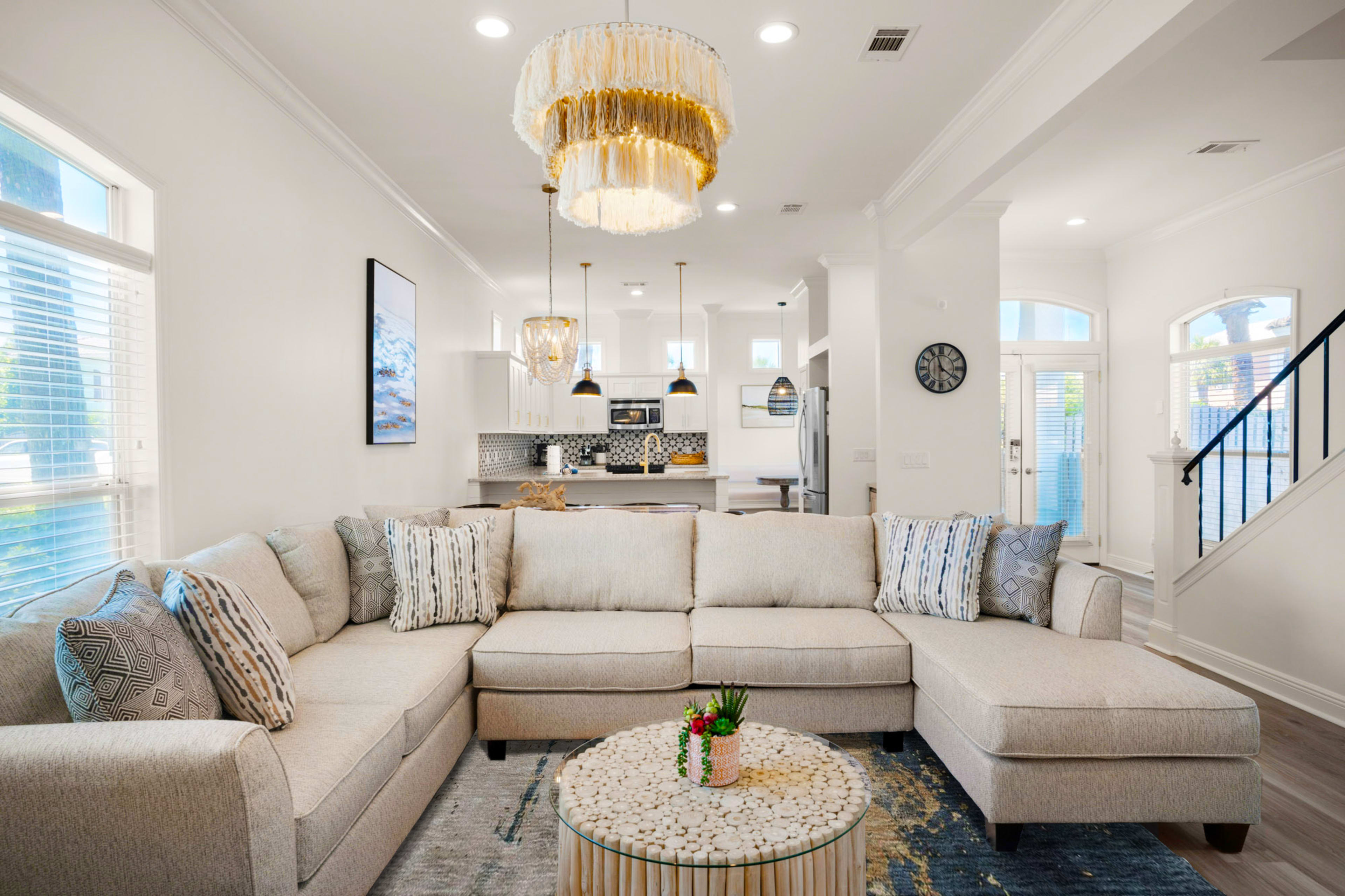 The image shows a modern living room featuring a large sectional sofa, a circular coffee table, and a well-lit open layout leading to a kitchen in the background.