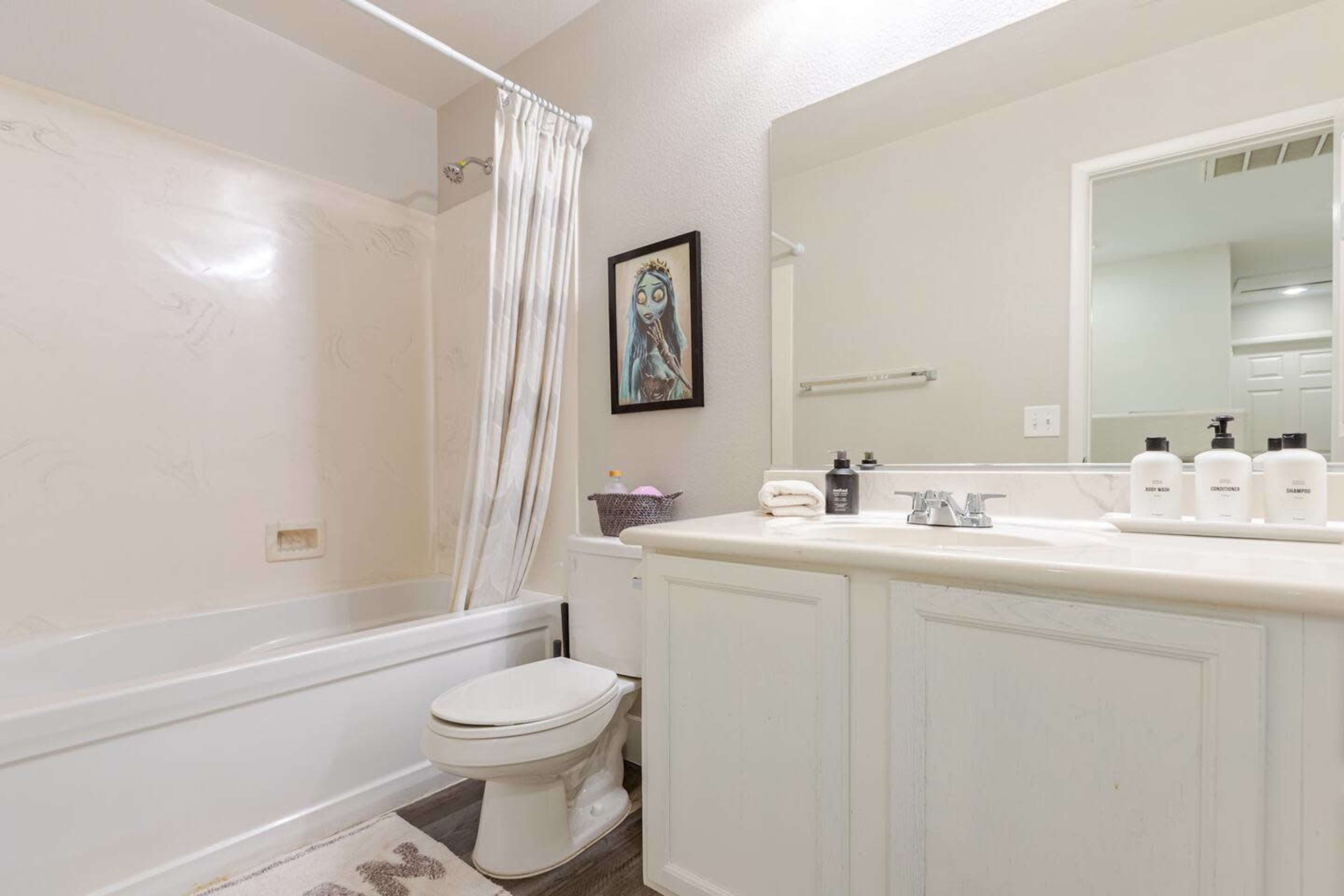 The image shows a bathroom featuring a bathtub, a toilet, a sink with several bottles lined up, and a framed artwork on the wall.
