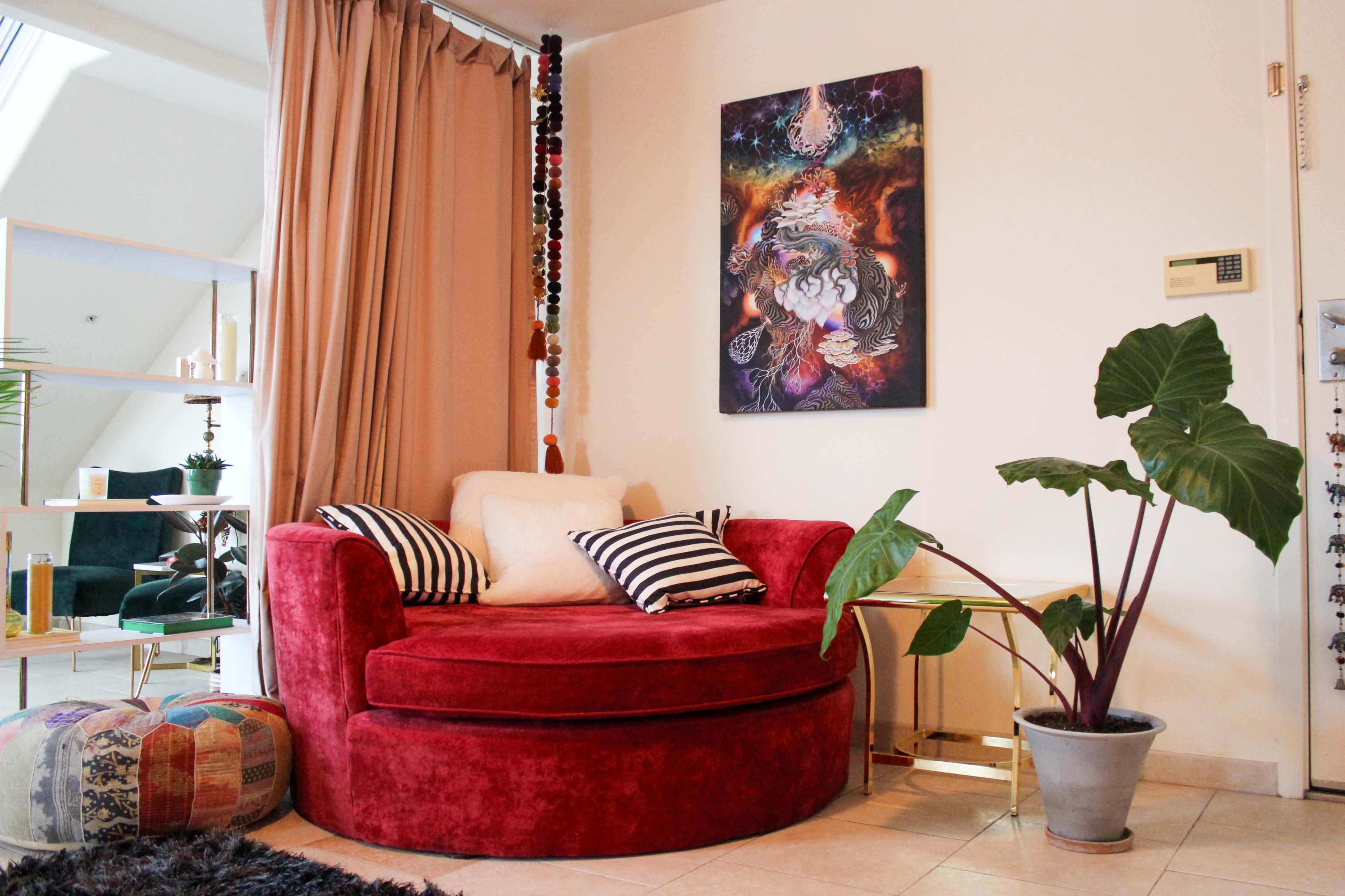 A cozy living space featuring a red circular sofa with pillows, a potted plant, and a colorful ottoman, all set against a wall adorned with a vibrant artwork.