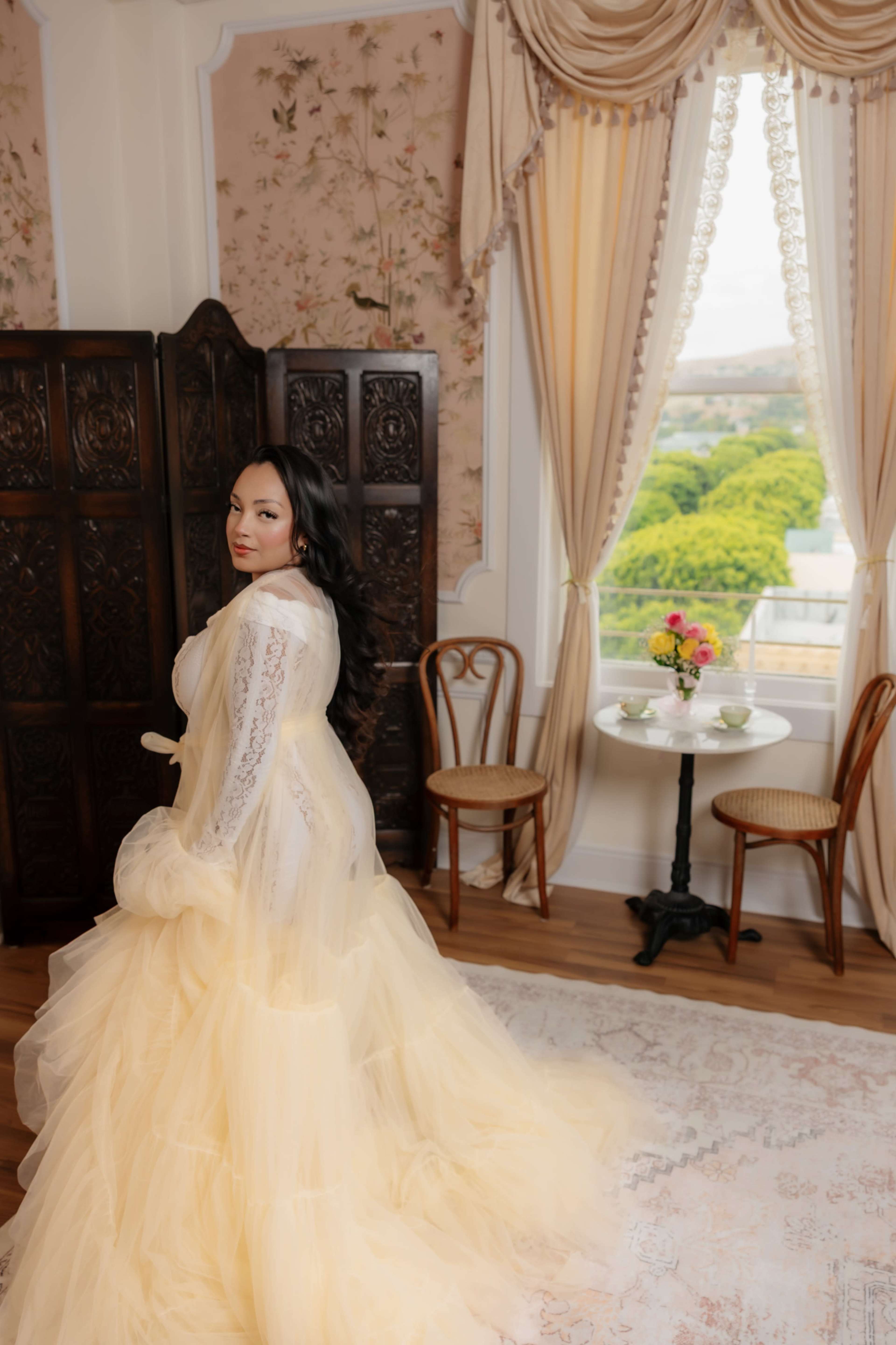 A woman in a yellow gown stands with her back turned, facing away from a window with a view of green trees, inside a beautifully decorated room.