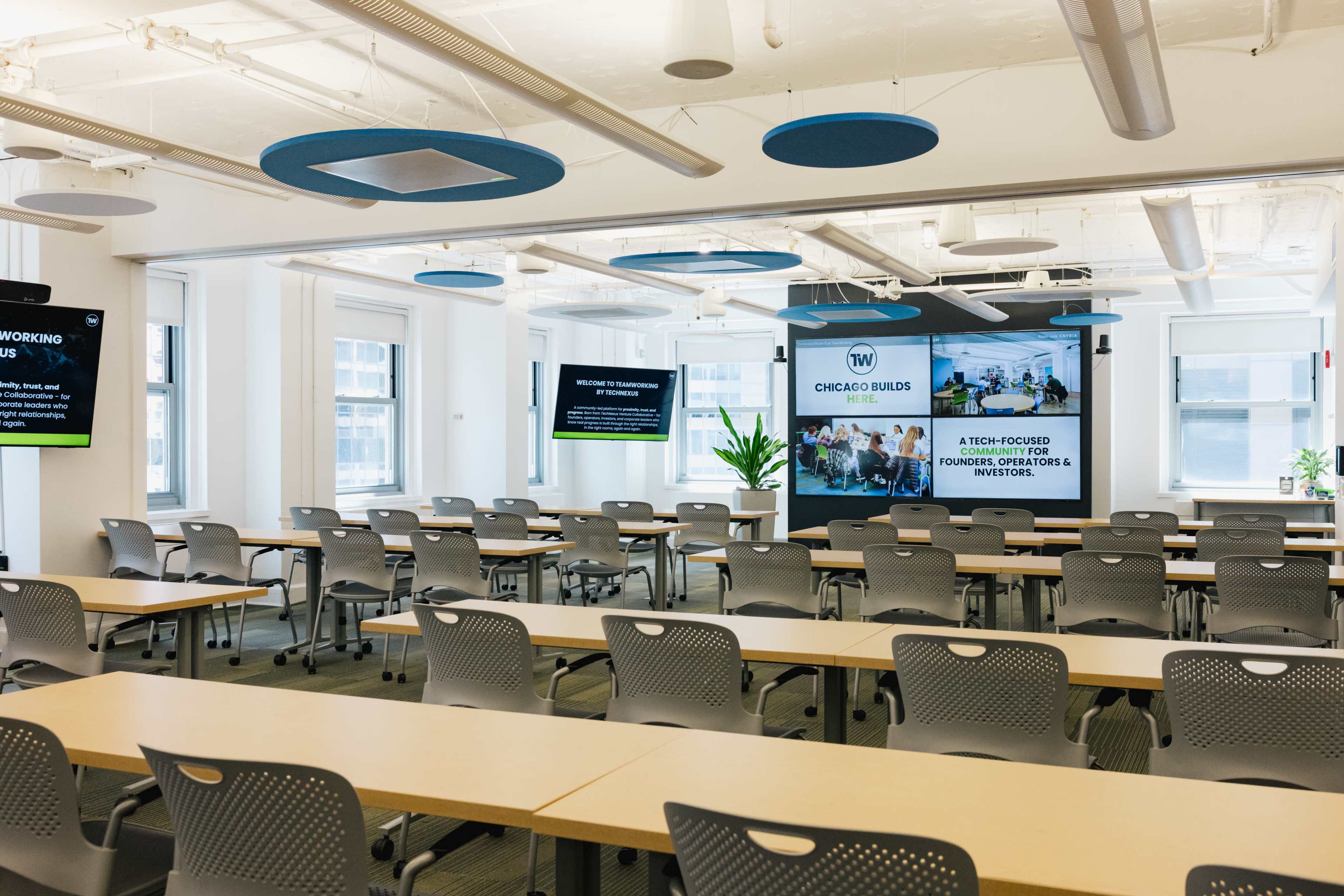 The image shows a modern conference room set up with rows of tables and chairs, featuring a large screen displaying information and a bright, open layout with natural light.
