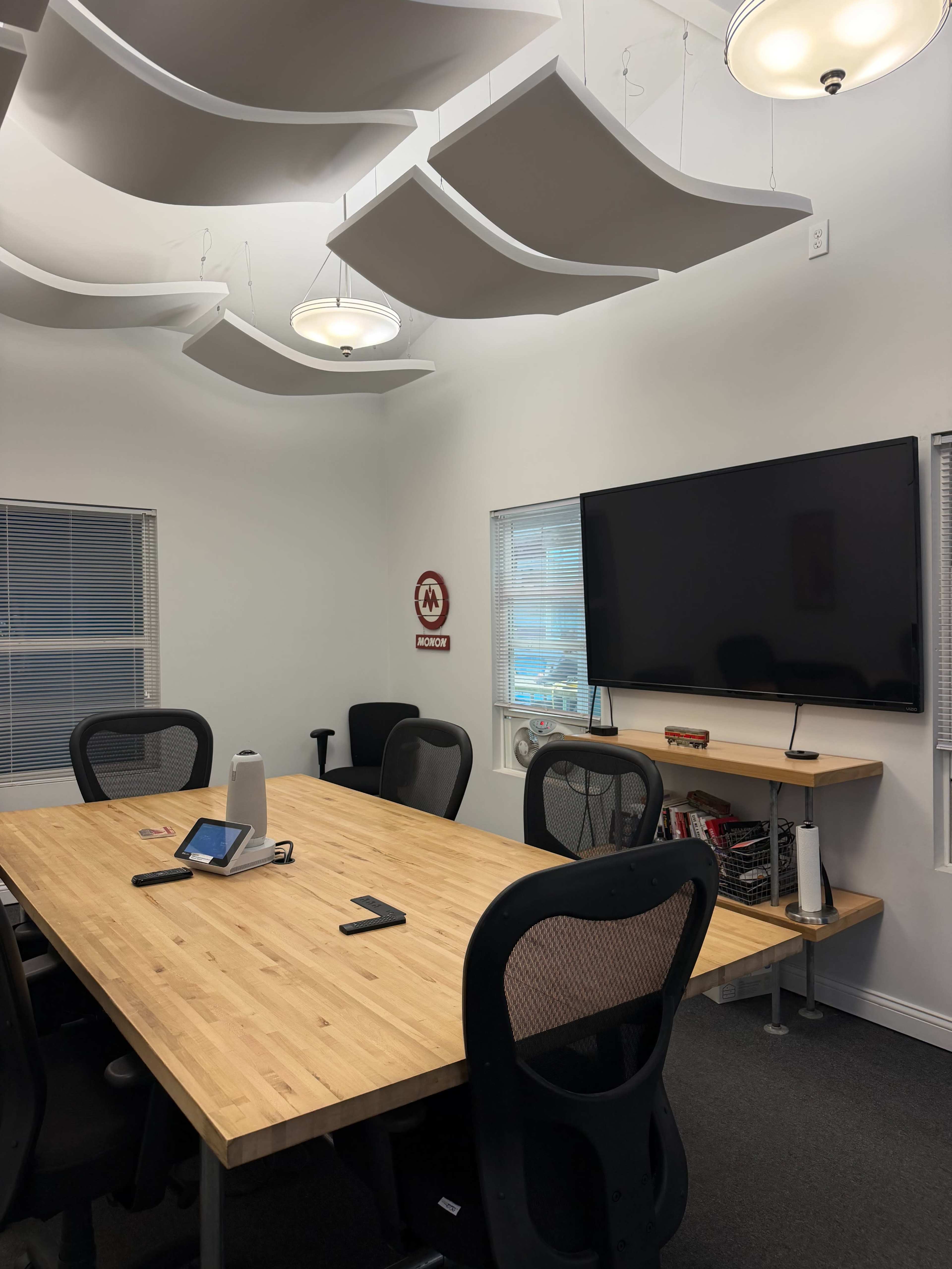 The image shows a modern conference room with a wooden table, black chairs, a large wall-mounted screen, and soft lighting from hanging fixtures.