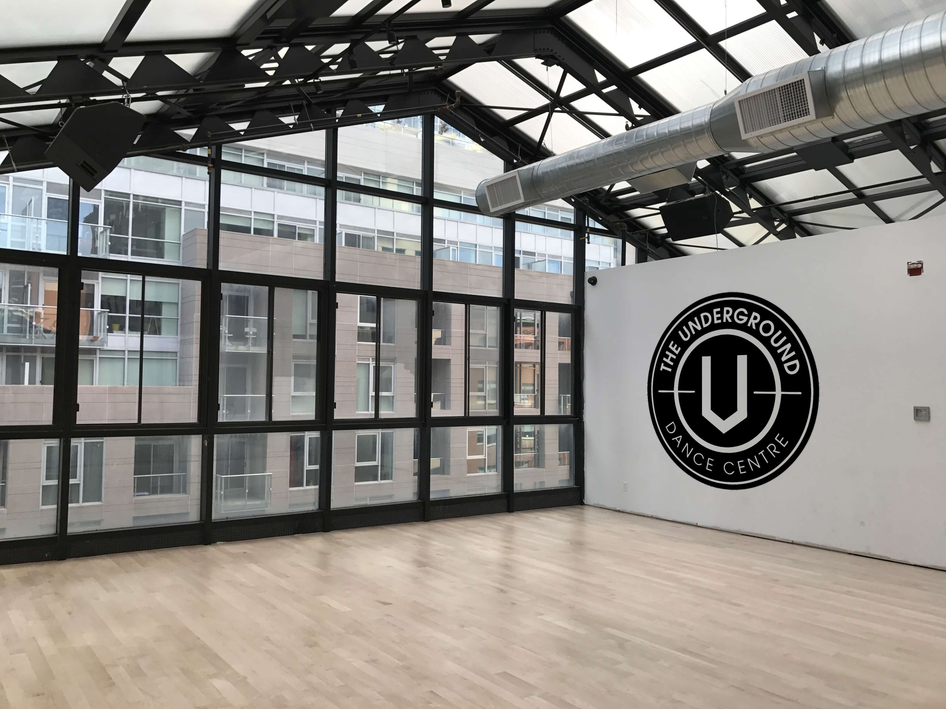 The image shows a large, empty dance studio with wooden flooring and floor-to-ceiling windows, featuring a logo for "The Underground Dance Centre" on one of the walls.