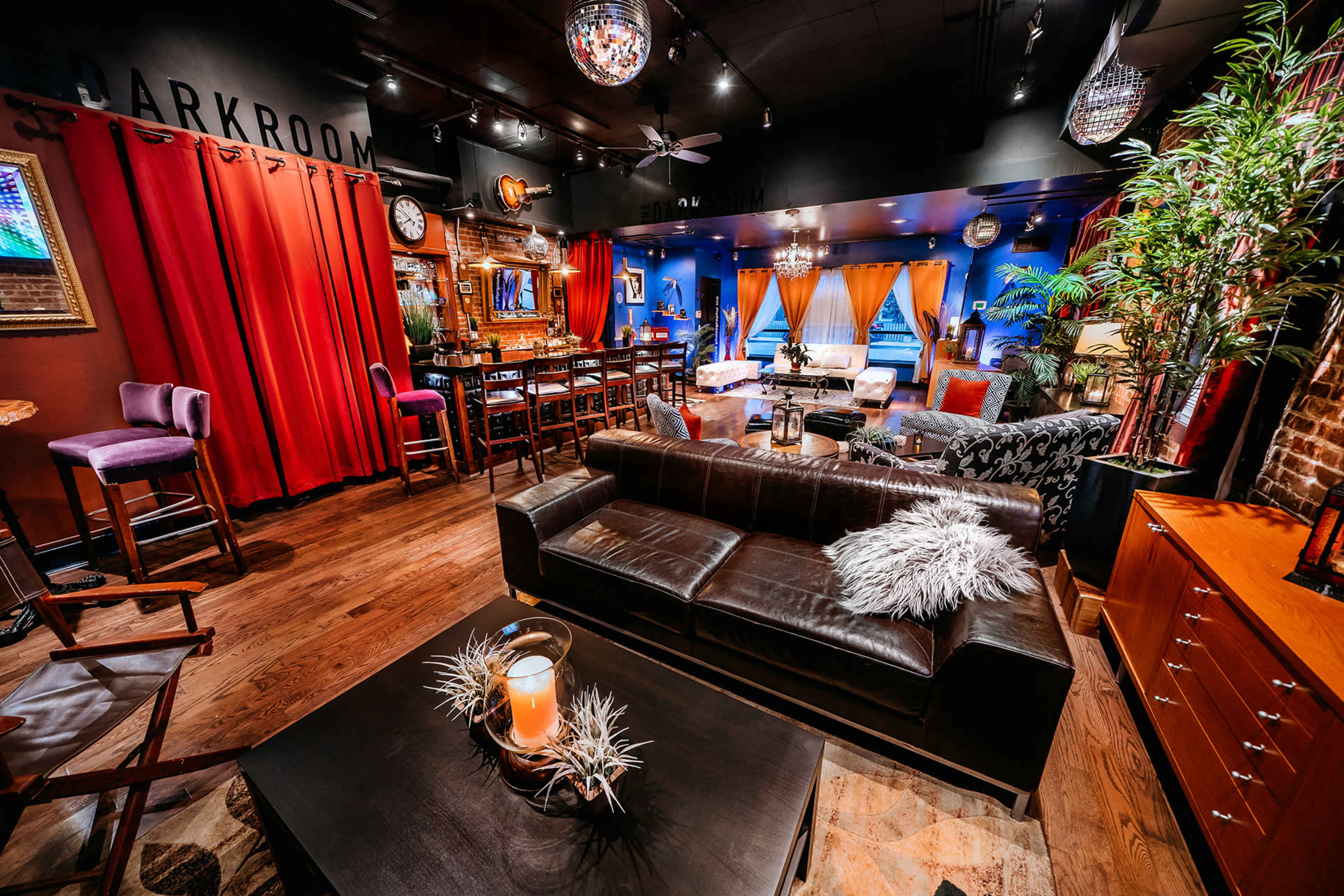 A stylish lounge with a leather sofa, bar area, and colorful curtains, illuminated by decorative lighting.