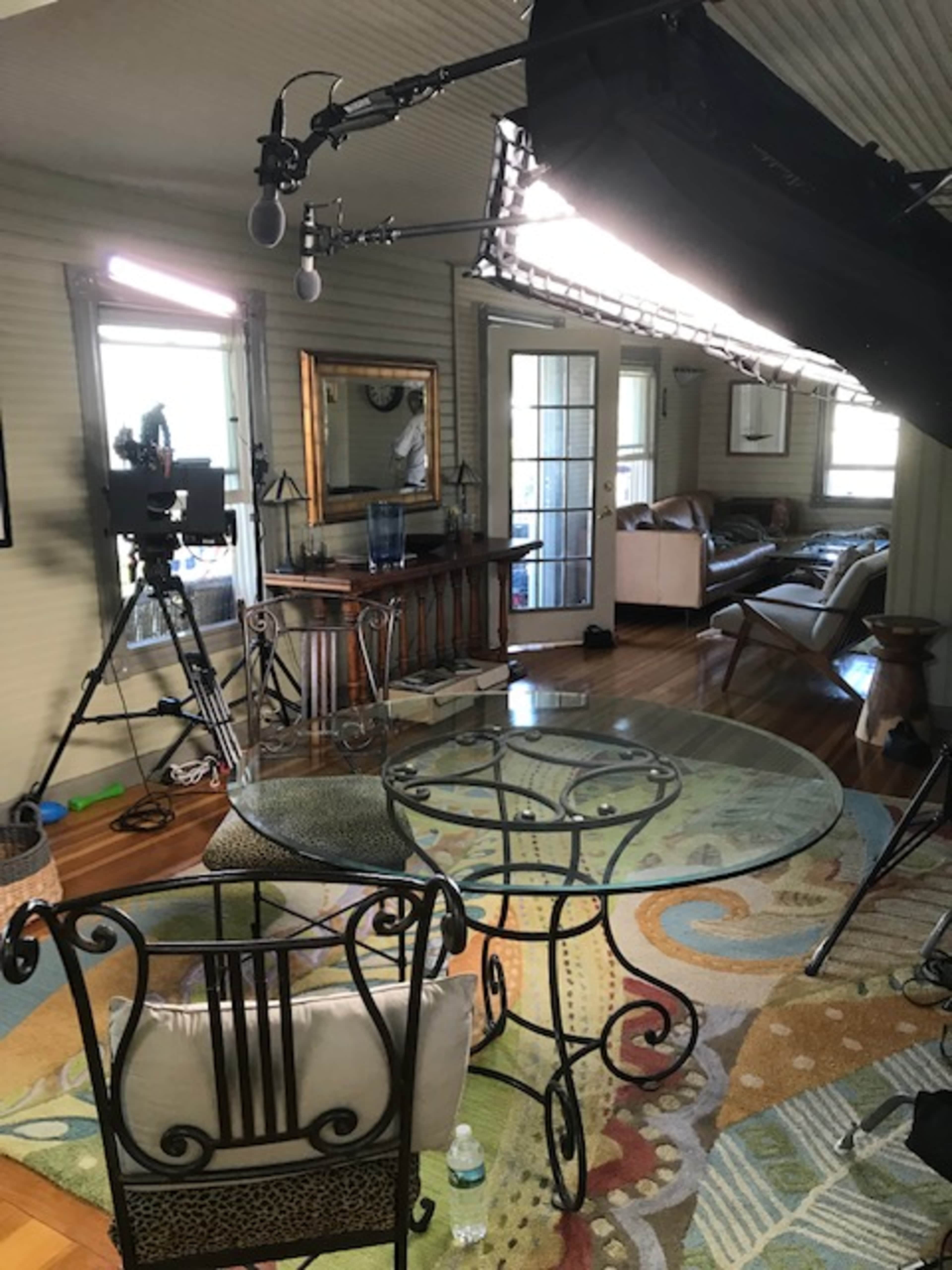 A living room setup for filming, featuring a glass-topped table with chairs, a camera on a tripod, and lighting equipment.