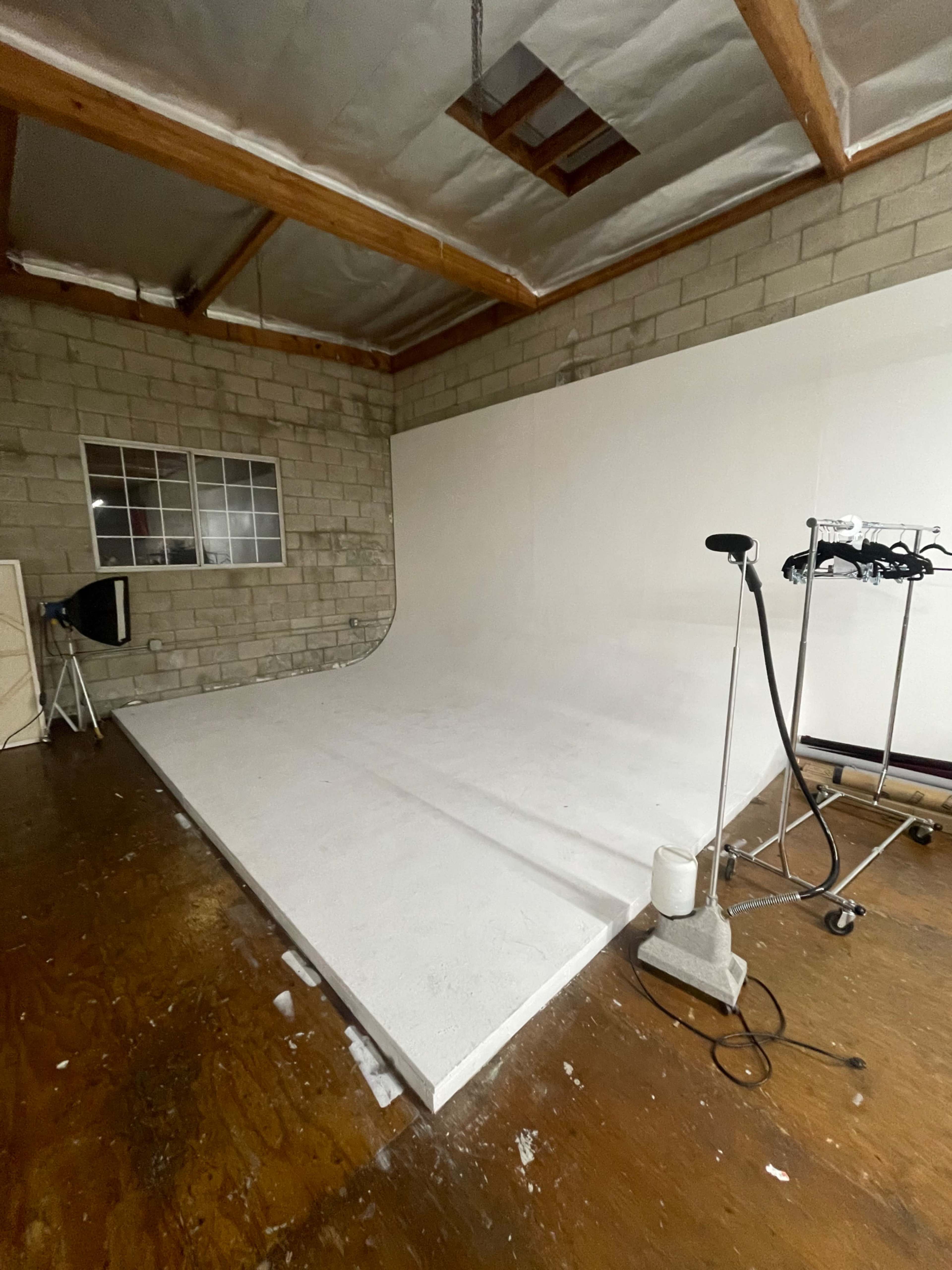 The image shows a sparsely furnished photography studio with a white backdrop and wooden flooring.