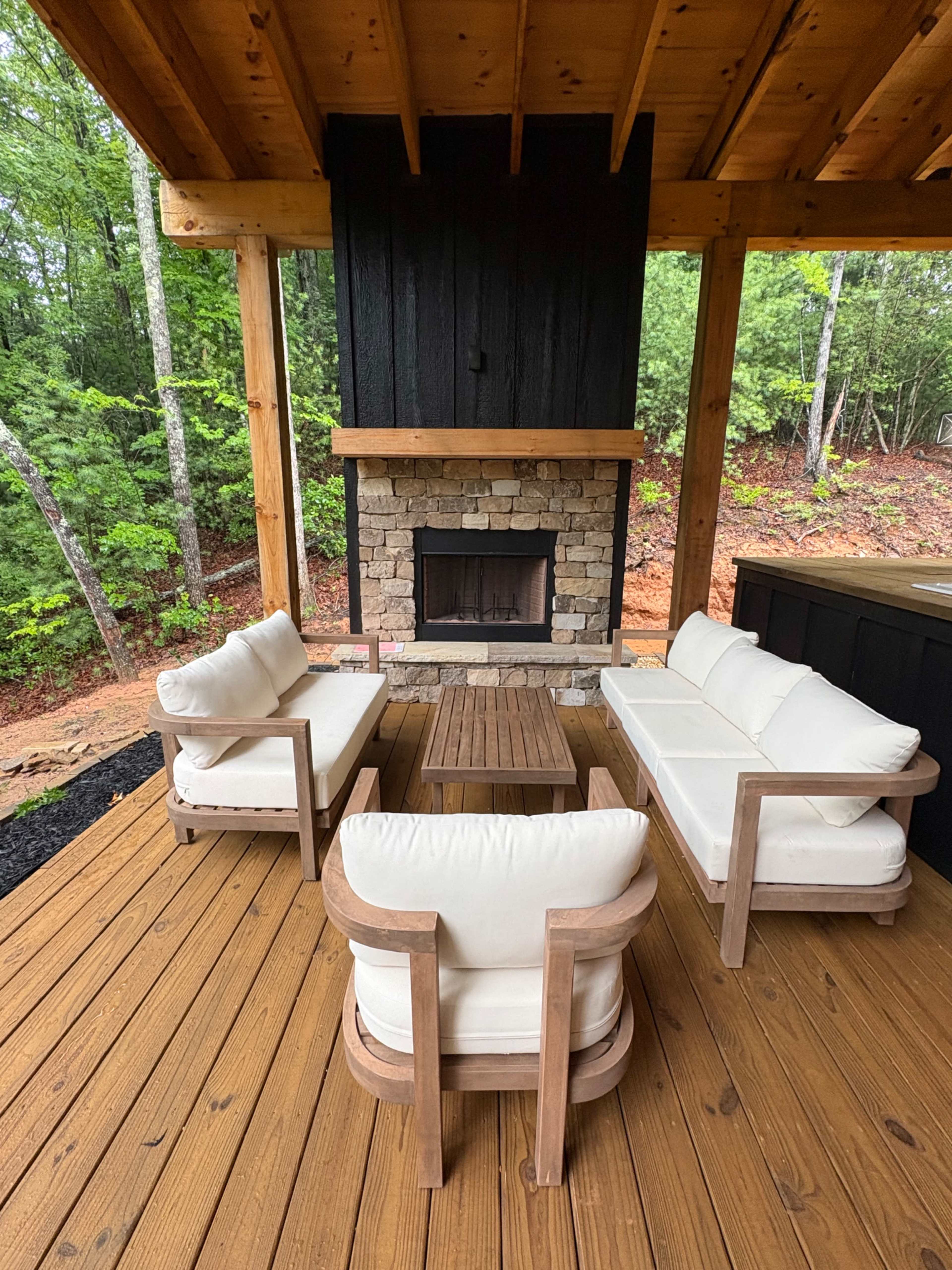 The image shows a cozy outdoor seating area featuring two white cushioned sofas and a coffee table arranged around a stone fireplace under a wooden structure surrounded by trees.