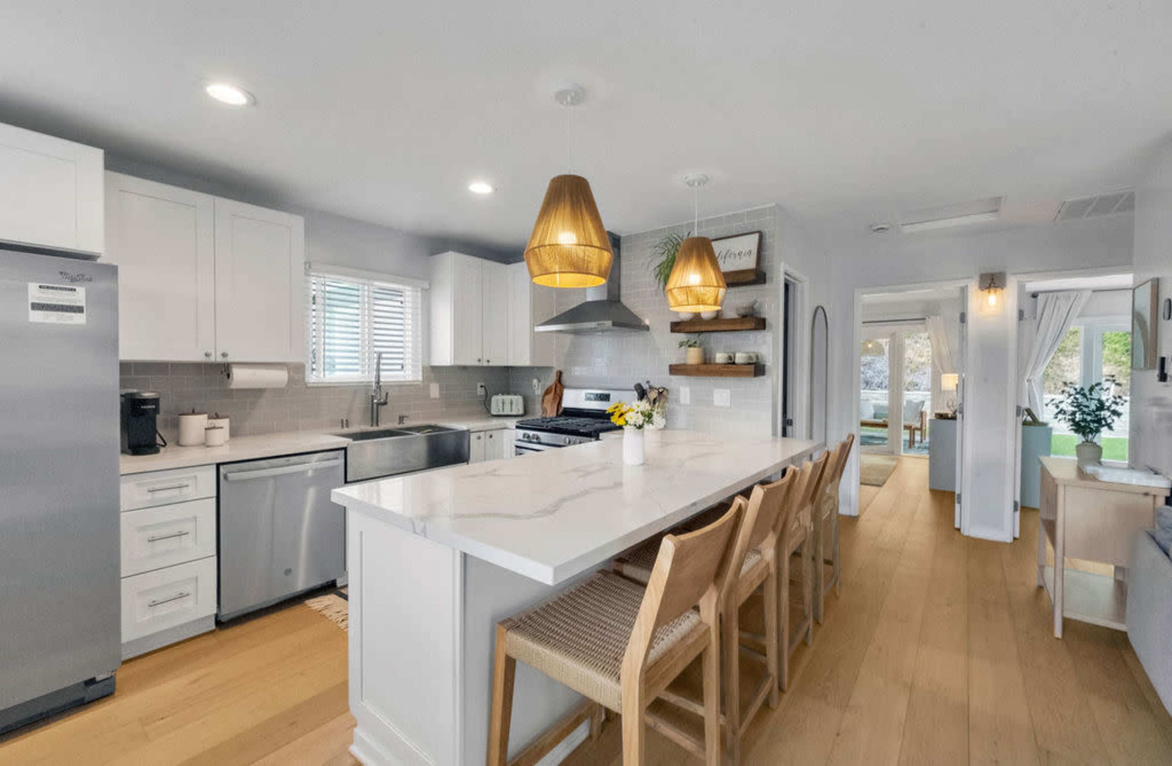 The image shows a modern kitchen with white cabinets, a large island with bar stools, and pendant lights, featuring stainless steel appliances and a dining area visible in the background.