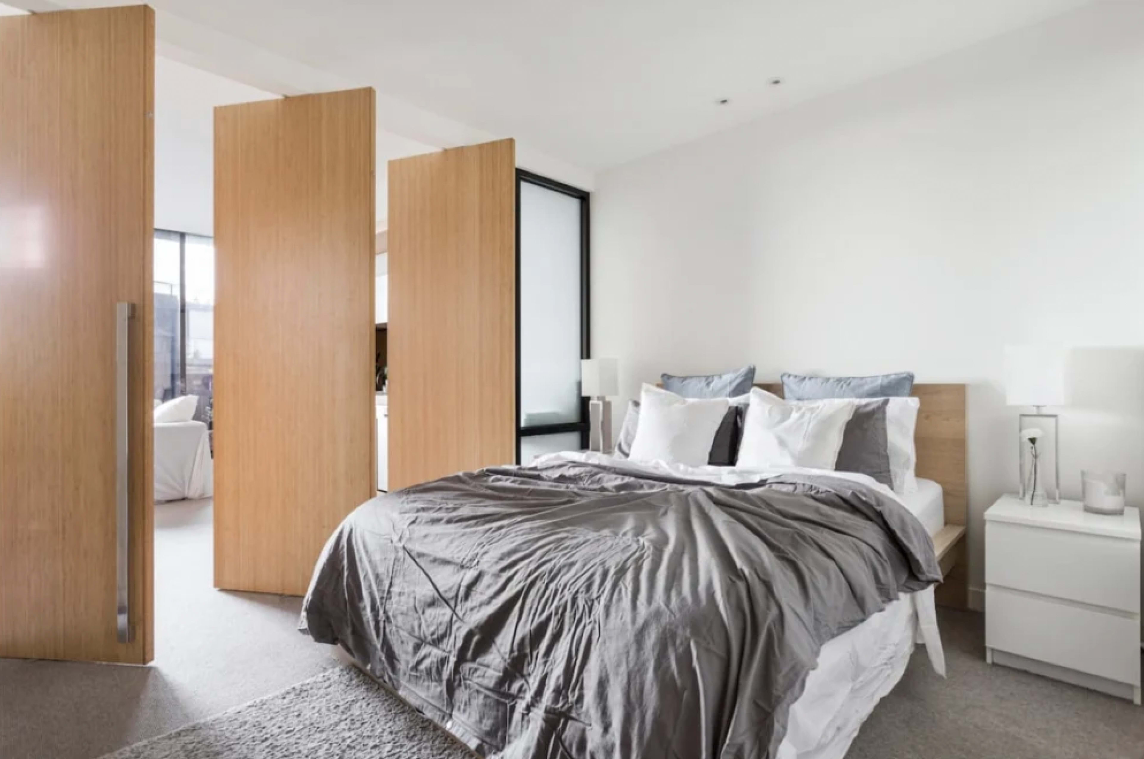 A modern bedroom features a bed with a gray comforter and white pillows, bordered by wooden room dividers and sunlight filtering through large windows.