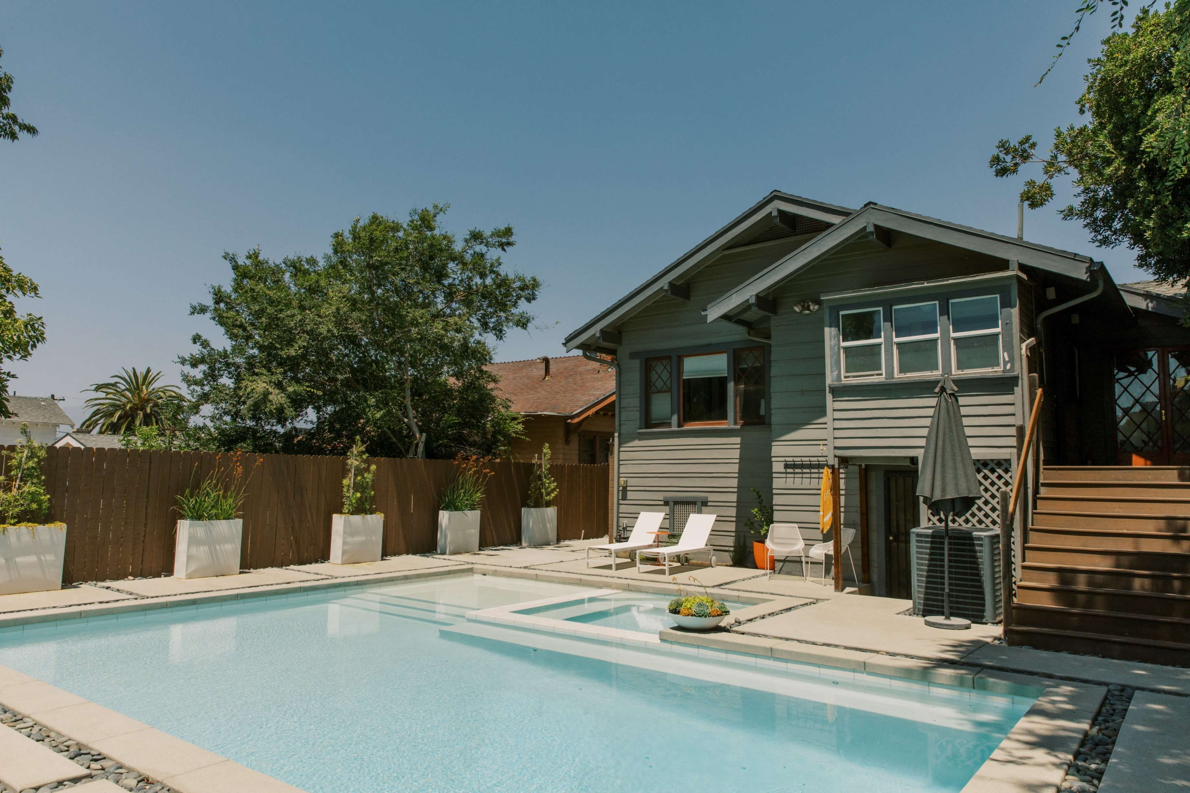 A modern gray house with multiple windows overlooks a rectangular swimming pool surrounded by potted plants and two lounge chairs.