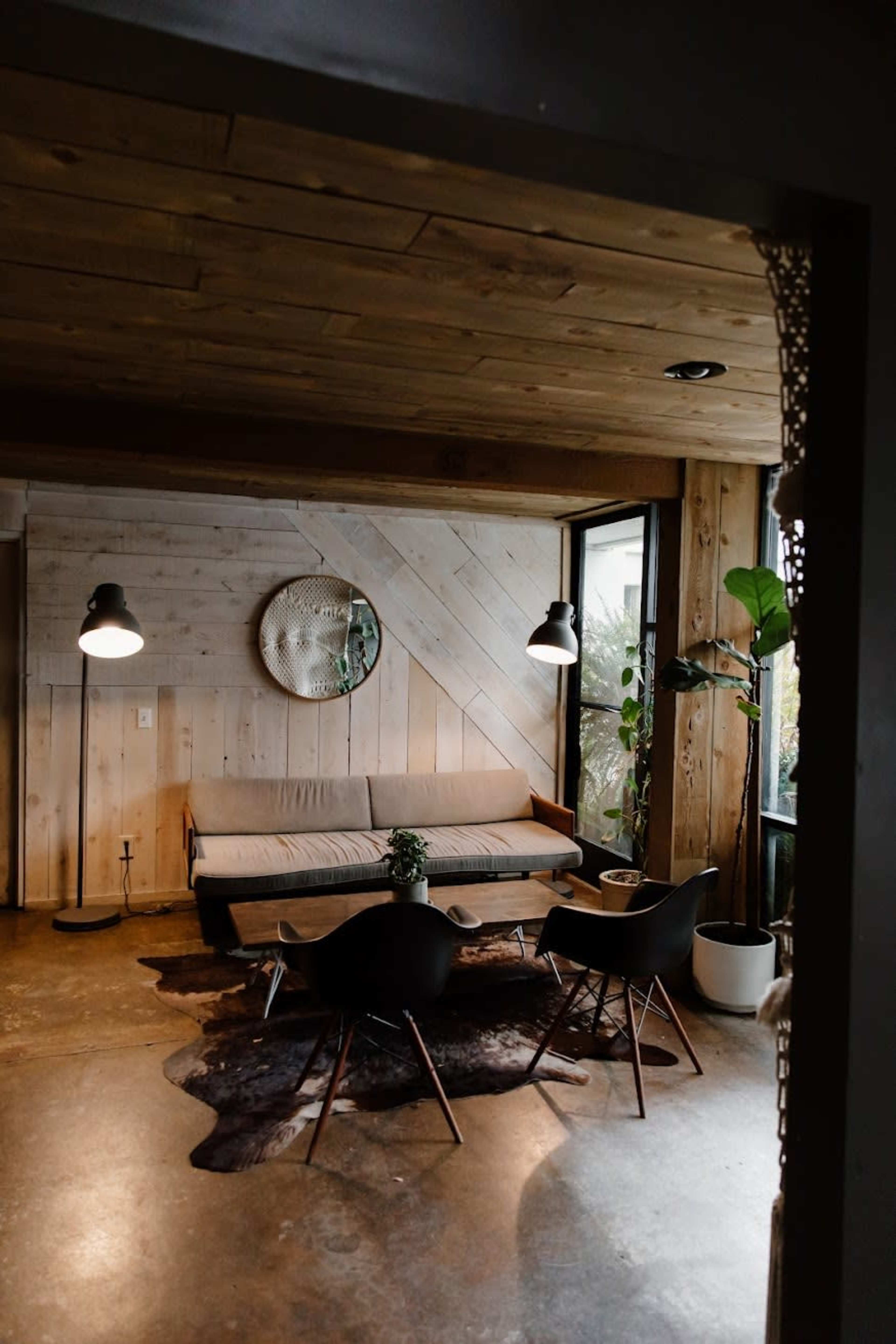 The image shows a modern living room with wooden walls, a sofa, a round mirror, and seating arranged around a cowhide rug.