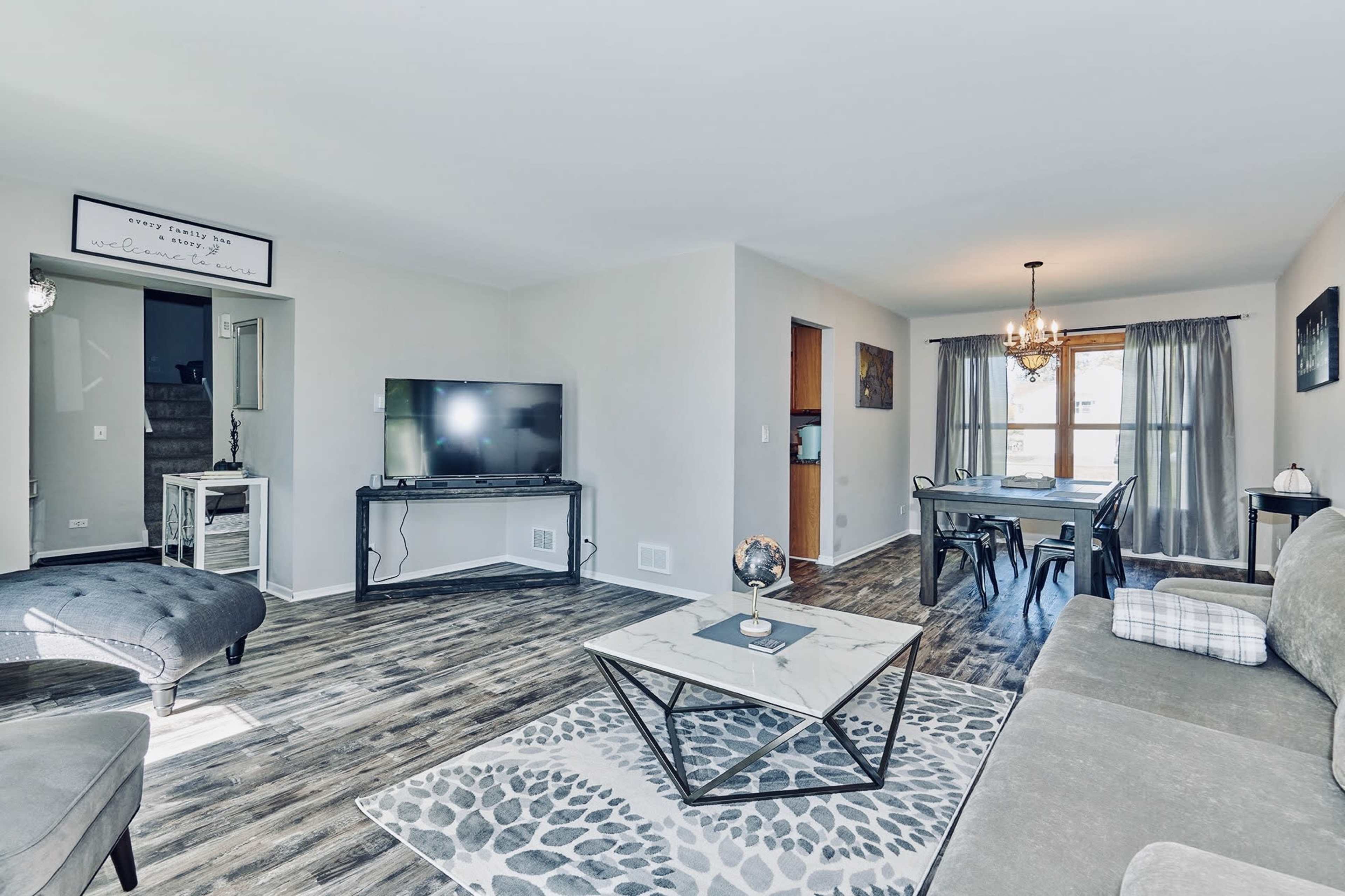 A modern living room featuring a gray sofa, a decorative coffee table, a TV on a stand, and a dining area with a chandelier.