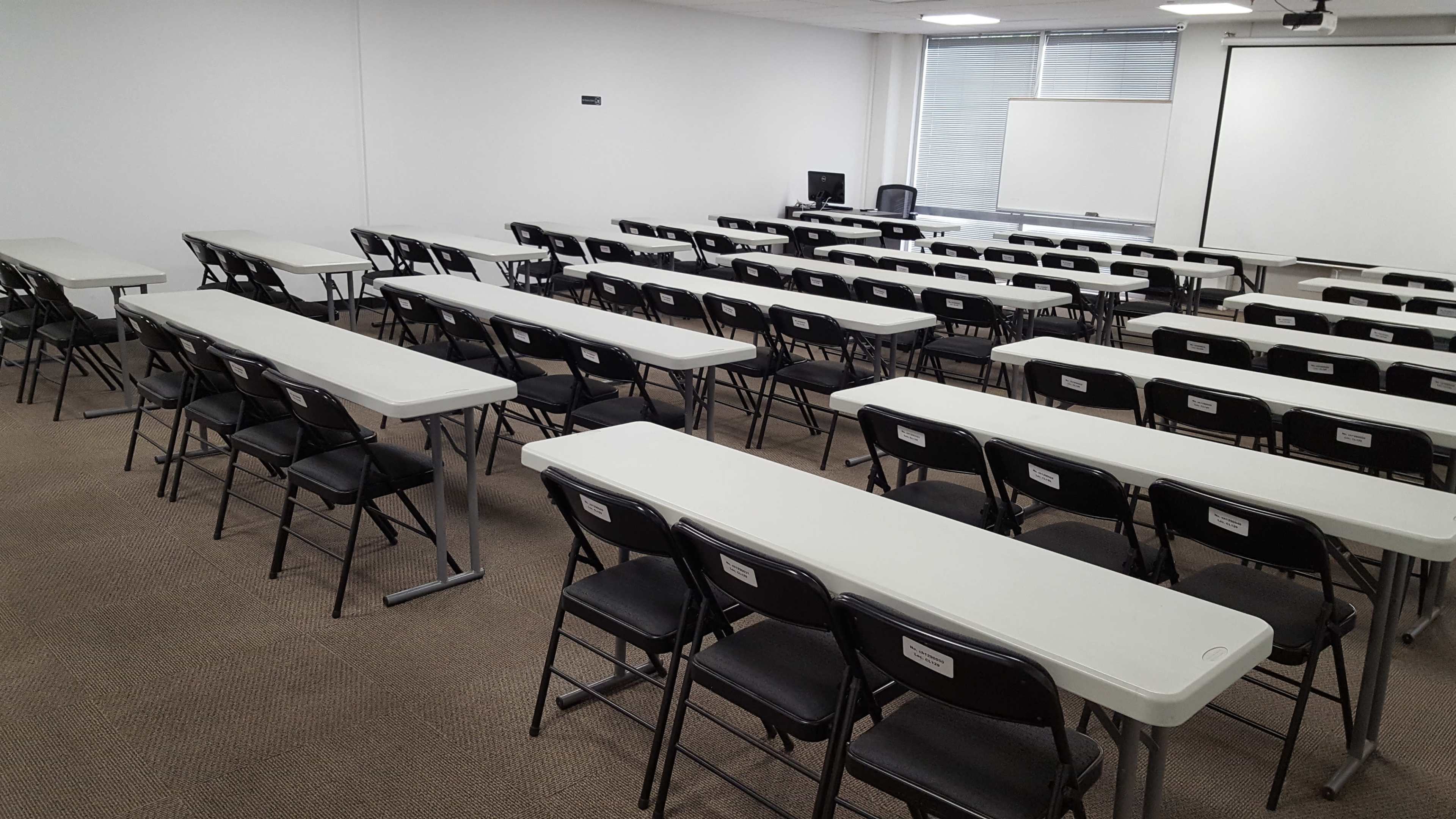 A large, empty classroom arranged with rows of white tables and black chairs.