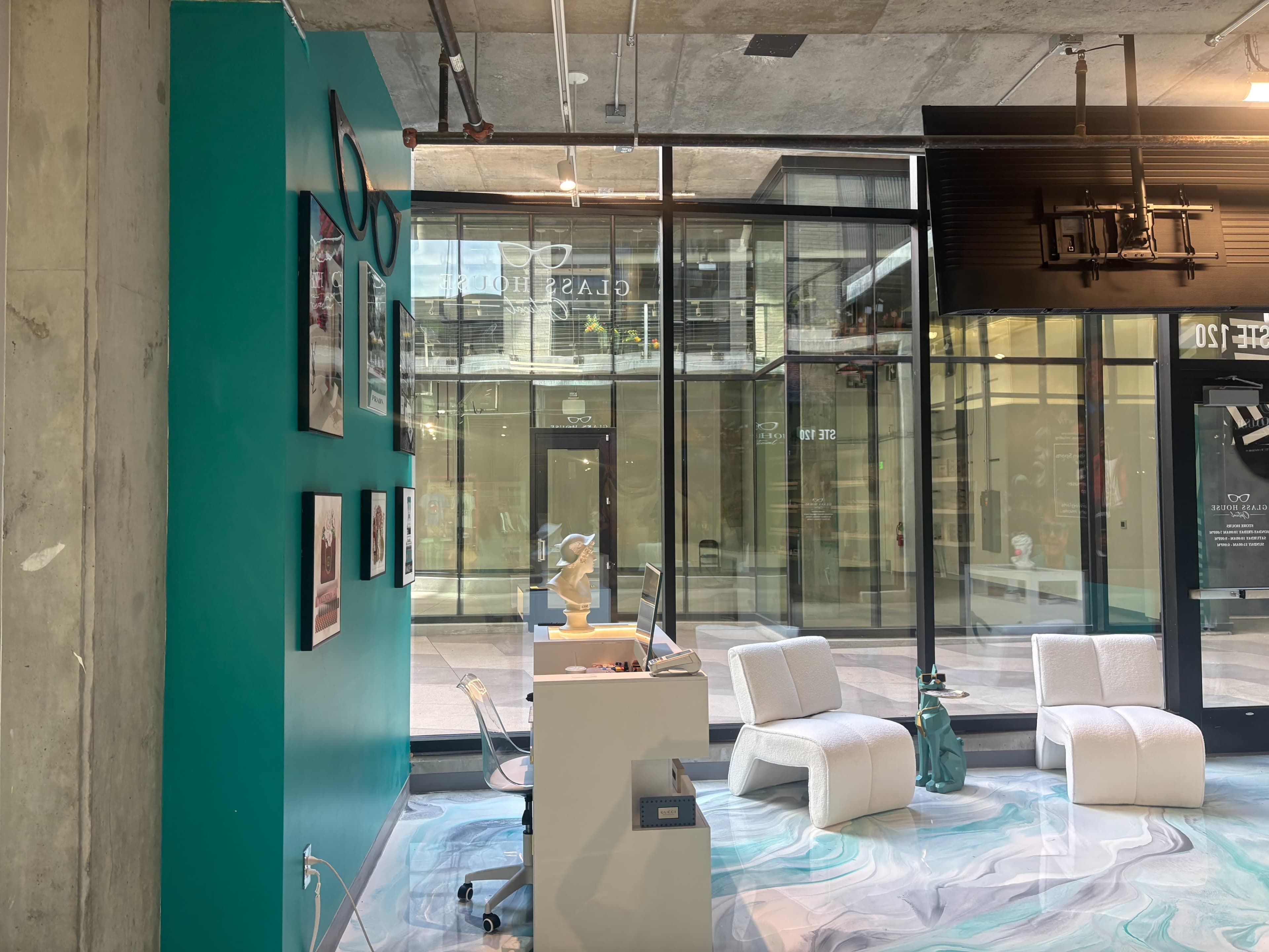 The image shows a modern interior space with a teal wall, displaying framed photographs, and a white desk accompanied by two white chairs on a marbled floor, overlooking a large glass storefront.