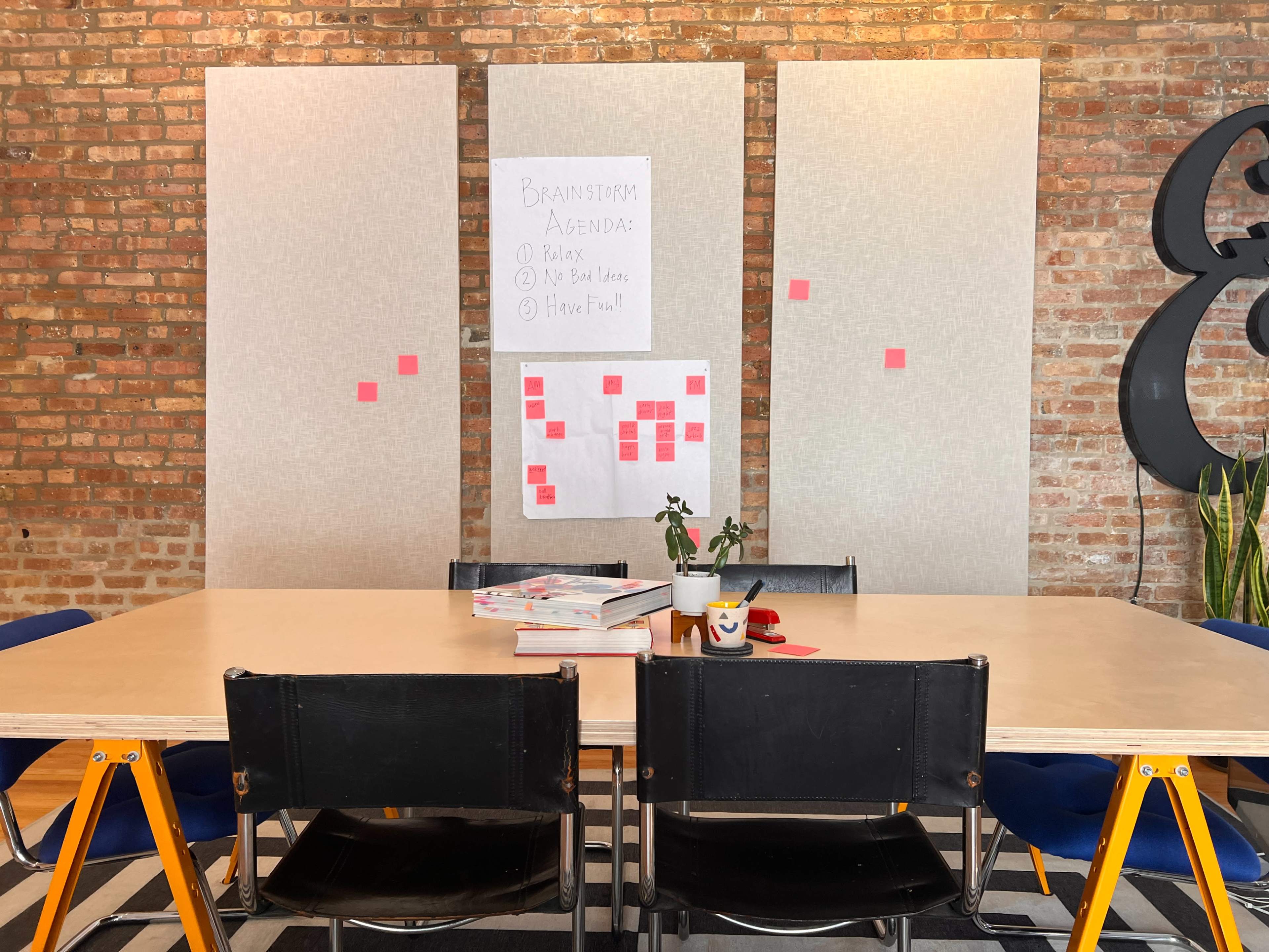 A modern meeting room features a long table surrounded by black chairs, with a whiteboard displaying a brainstorming agenda and post-it notes on the wall.