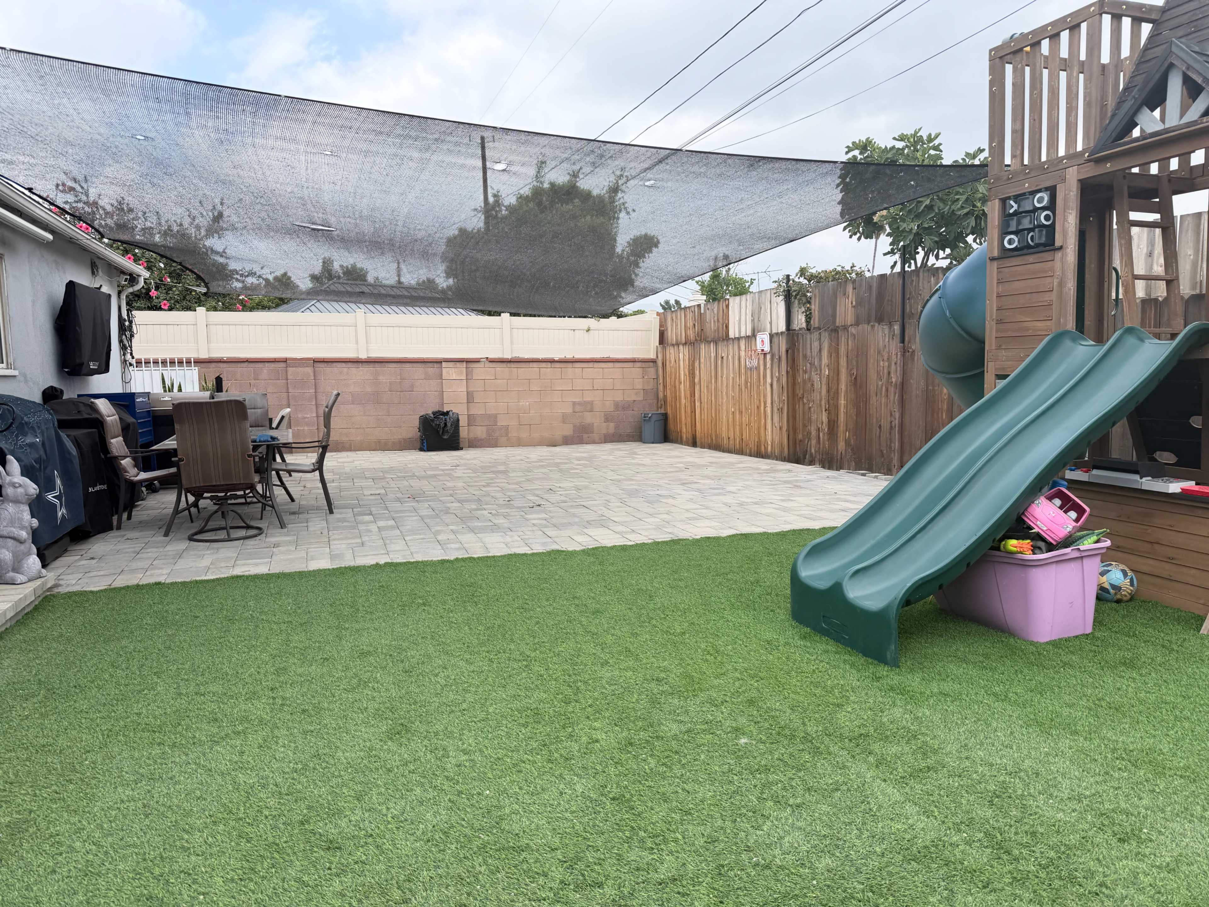 The image shows a backyard with synthetic grass, a wooden play structure with a slide, a dining table with chairs, and a shaded area overhead.