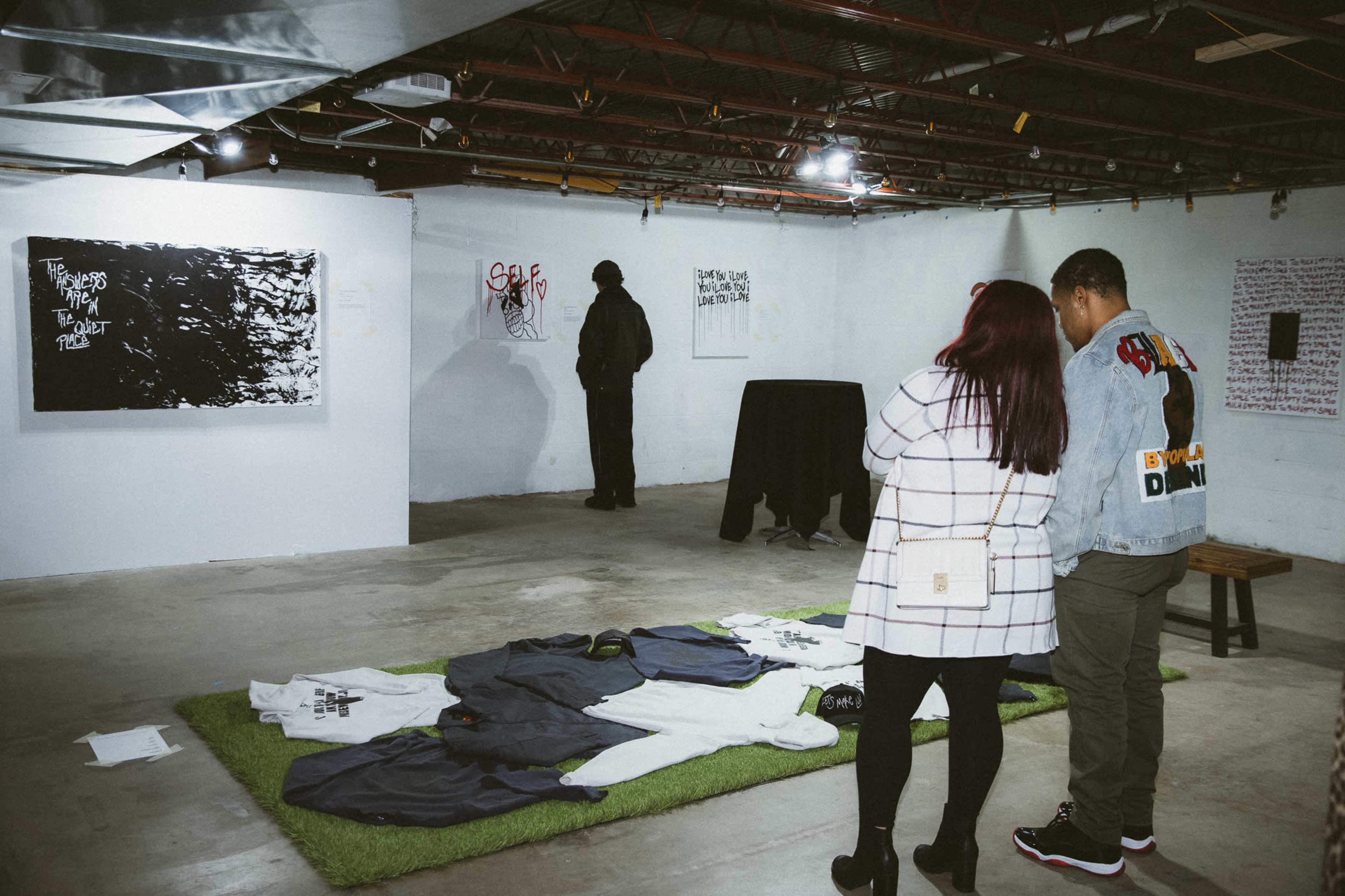 An art exhibition in a basement with two individuals examining artwork on the walls and clothing displayed on the floor.