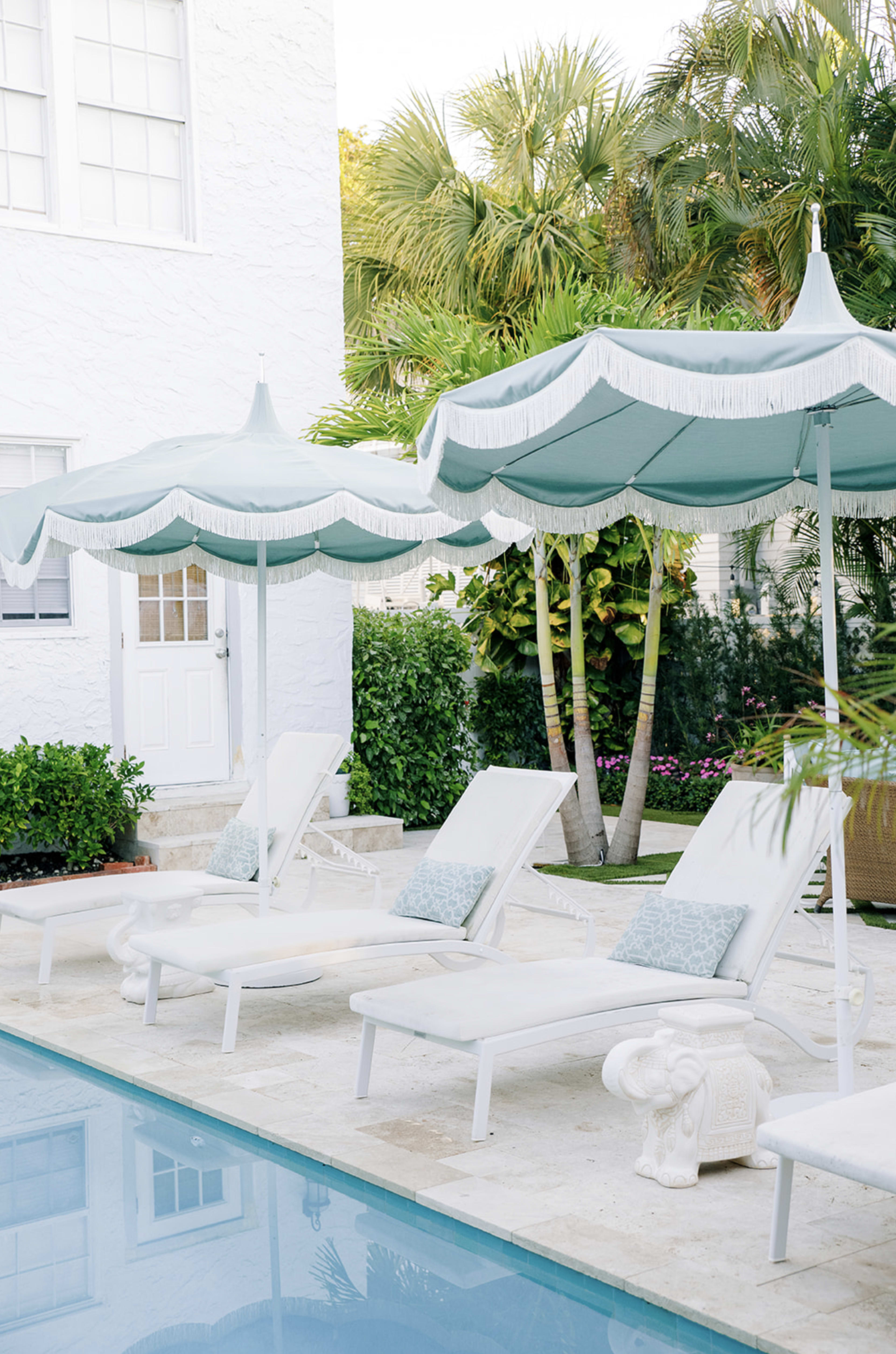 The image shows a serene pool area with white lounge chairs and teal-striped umbrellas surrounded by lush greenery.