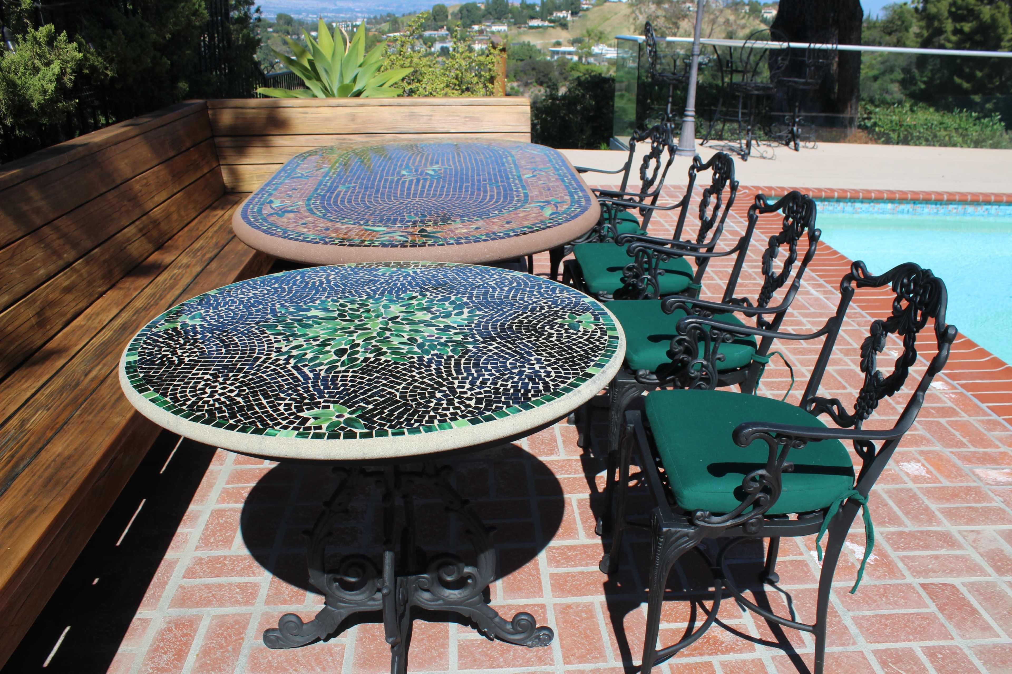 The image shows two ornate mosaic-tiled tables accompanied by wrought iron chairs around a poolside setup.