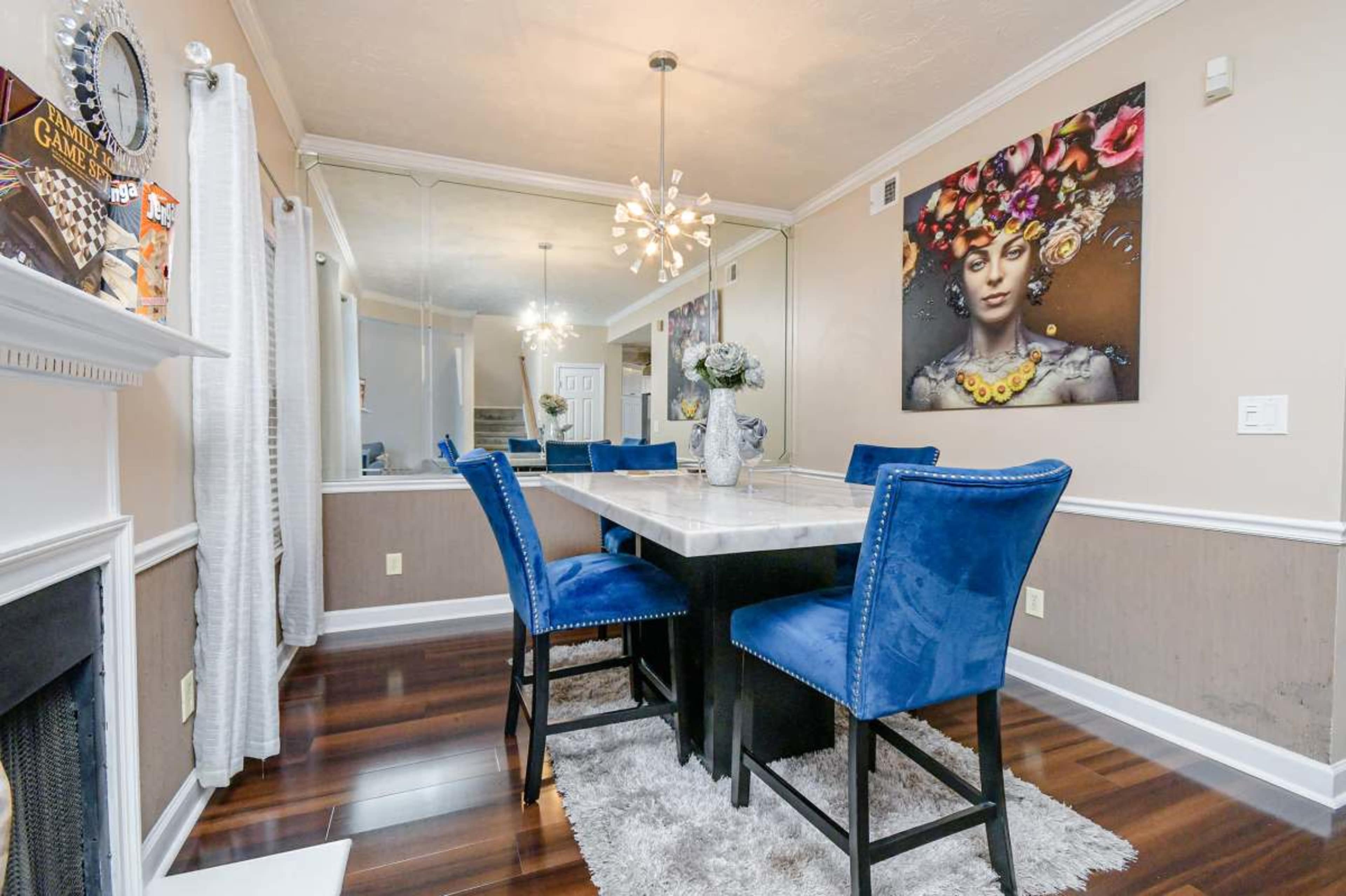 The image shows a dining area with a marble table surrounded by blue upholstered chairs, a decorative wall mirror, and a large artwork featuring a woman.