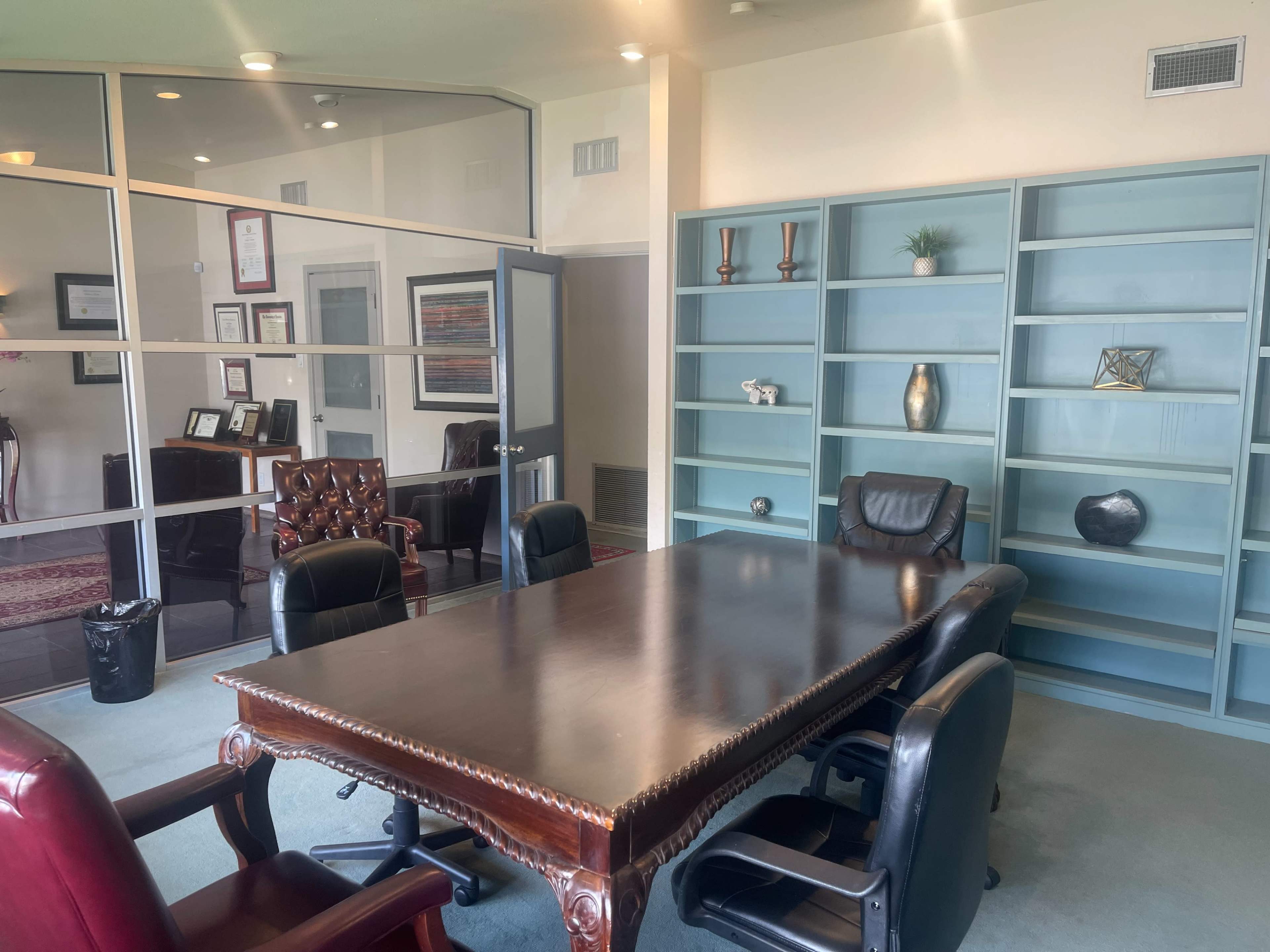 A modern conference room with a large wooden table surrounded by black leather chairs and a bookshelf filled with decorative items.