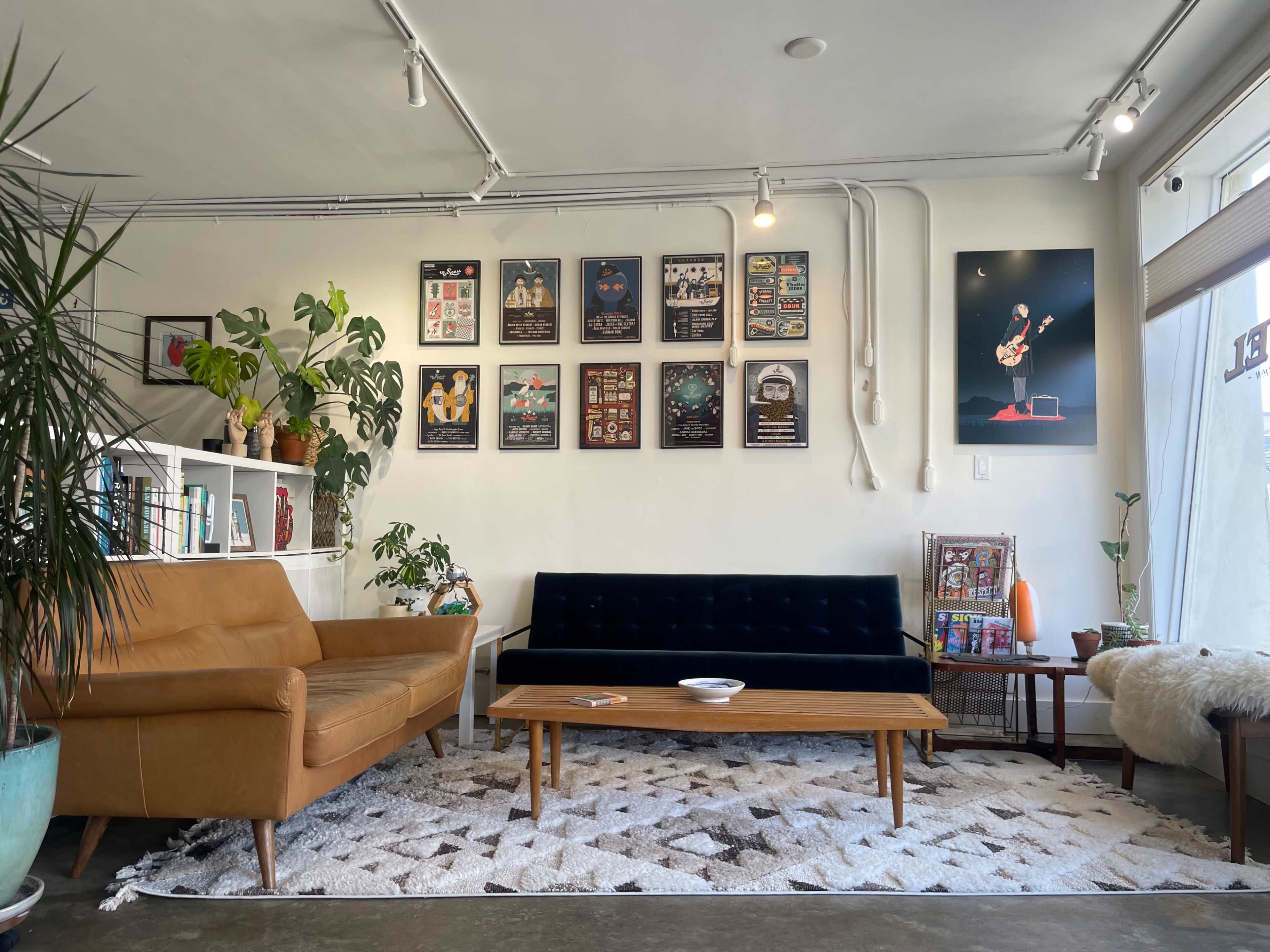 The image shows a modern living space featuring a dark blue sofa, a brown leather armchair, a wooden coffee table, and several framed posters on the wall, with plants and a decorative rug on the floor.