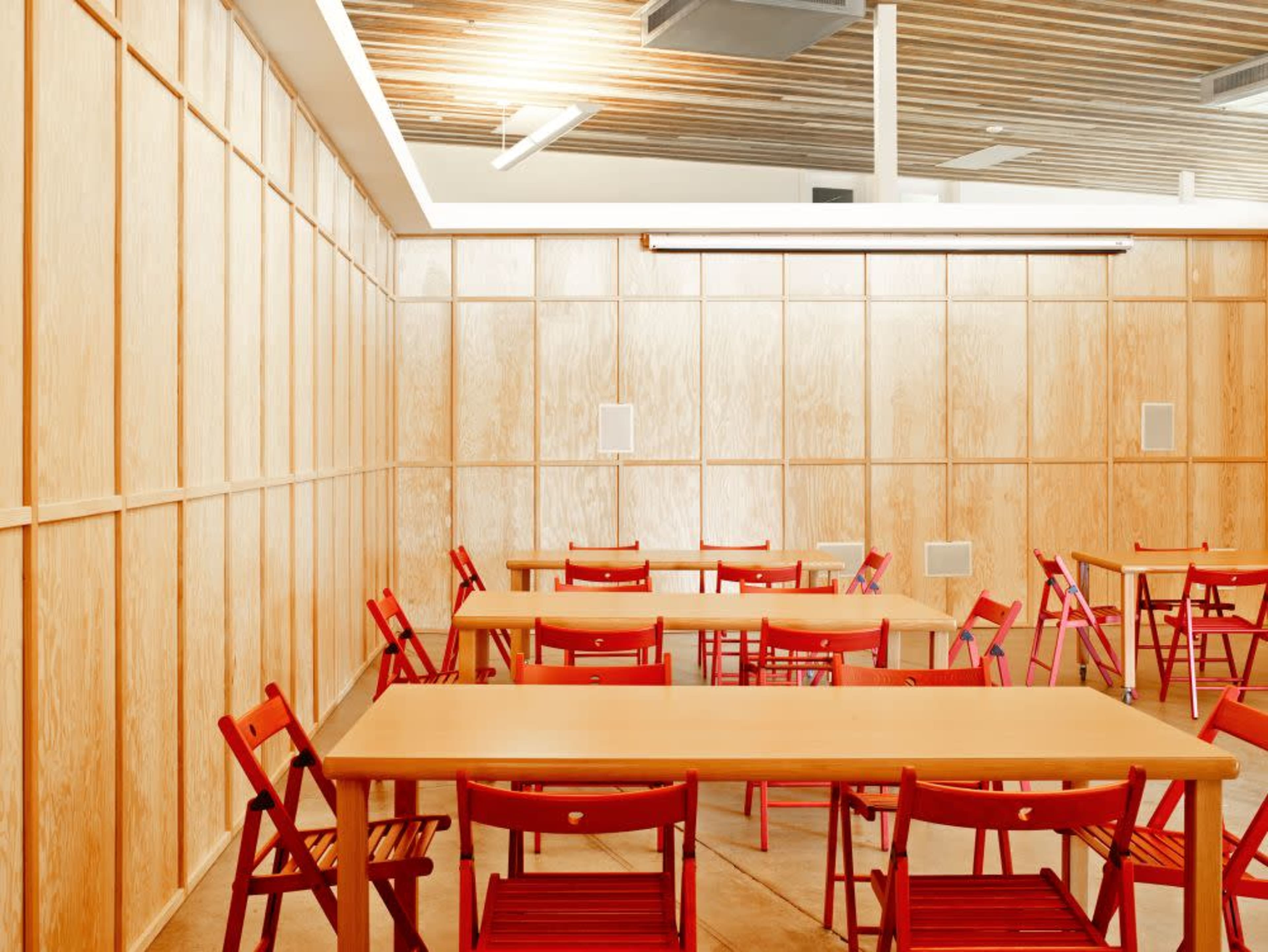The image shows a room with several tables and red chairs arranged neatly against wooden paneled walls.