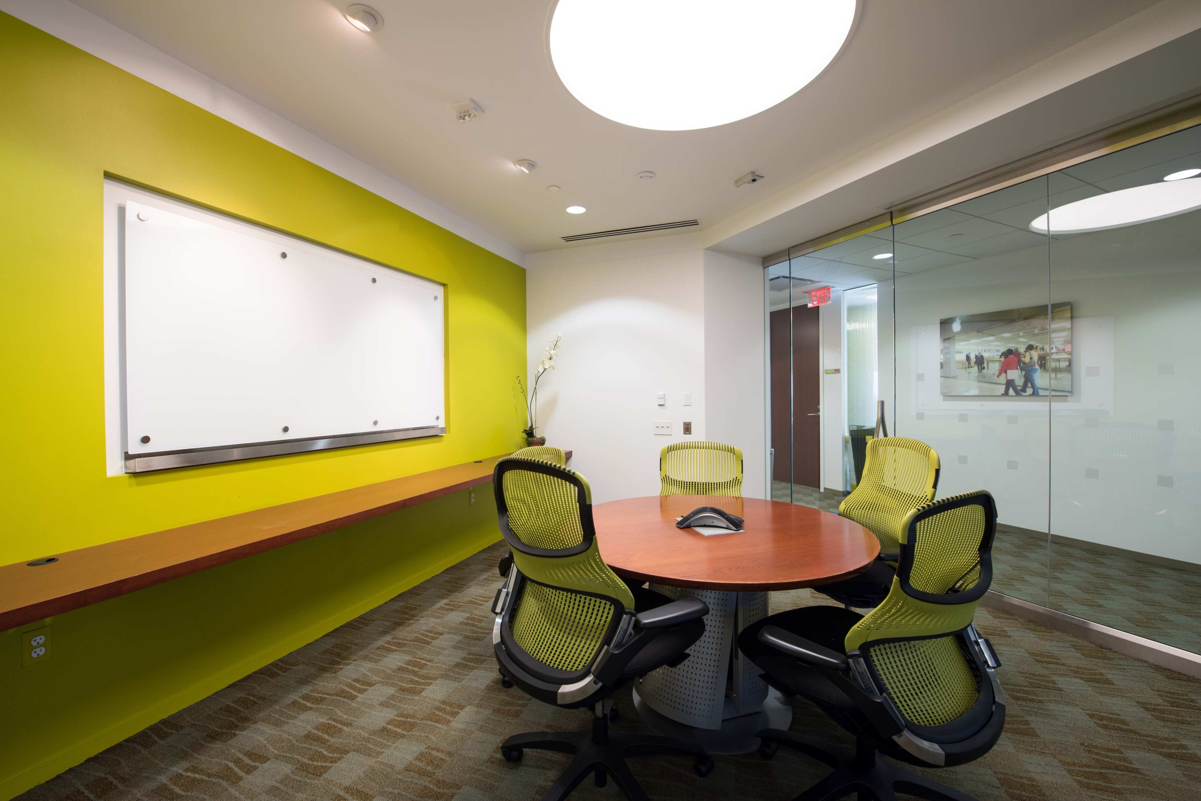 The image shows a modern conference room featuring a round table surrounded by four green chairs, a wall-mounted whiteboard, and a large circular ceiling light, with a bright green wall and glass-paneled entrance.
