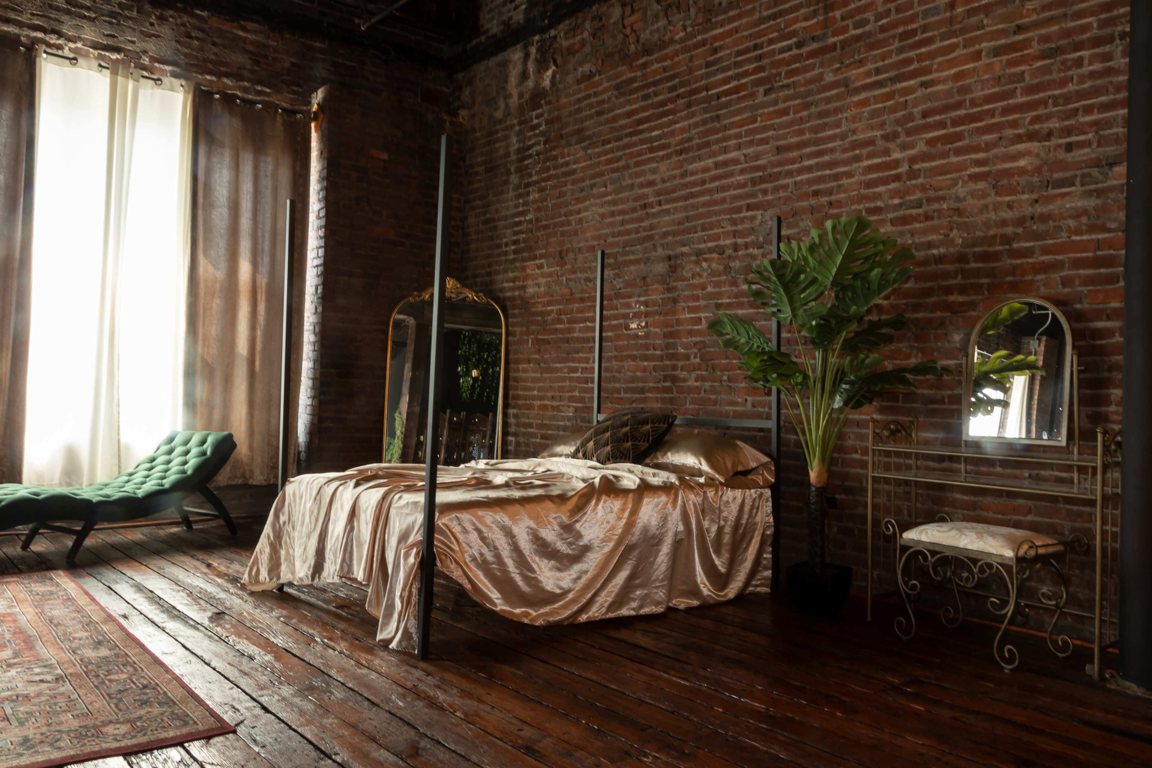 The room features a bed with satin bedding, a vintage mirror, a potted plant, and large windows alongside exposed brick walls.