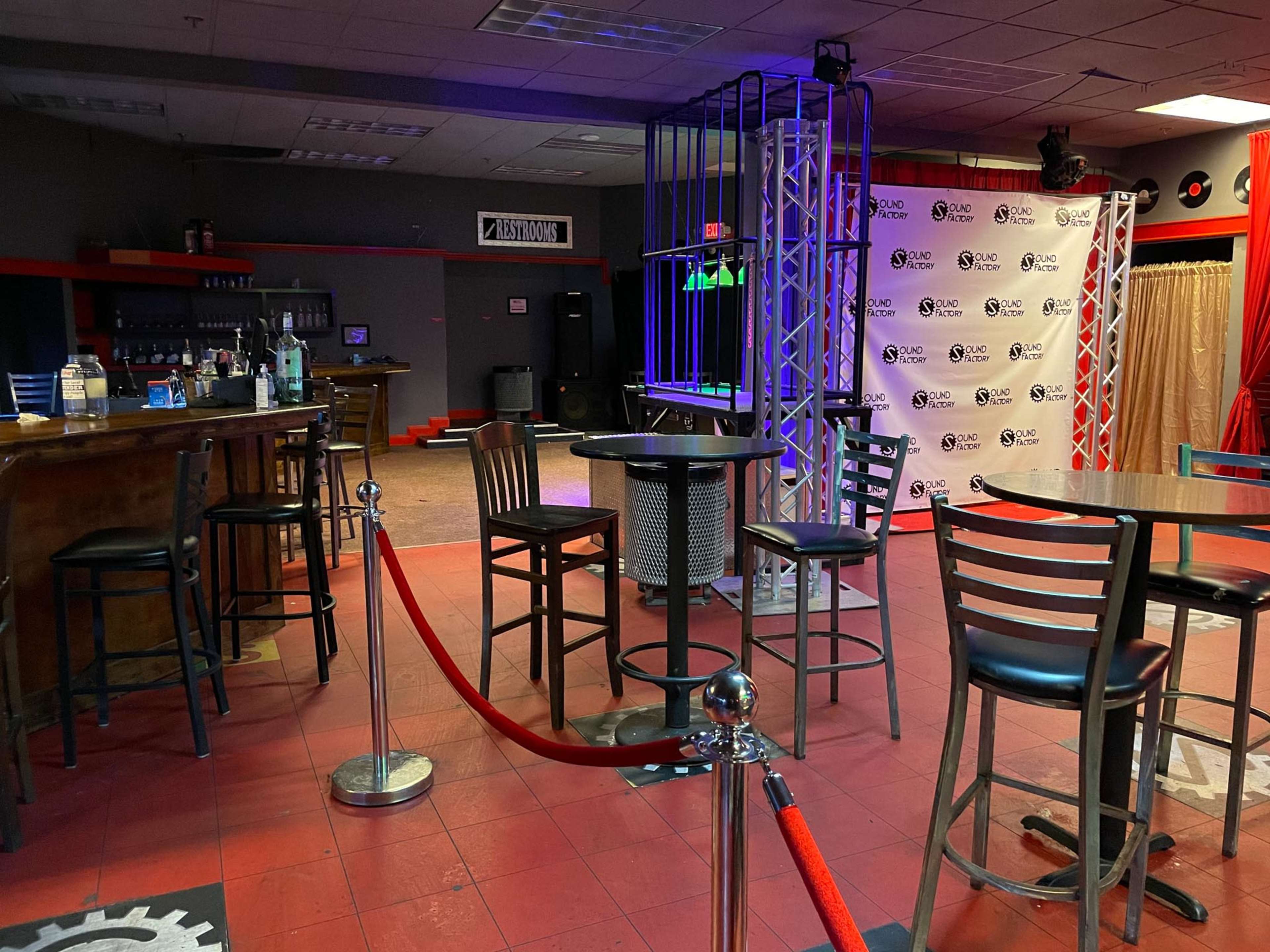 A dimly lit bar area with high tables, stools, and a decorative backdrop featuring a logo.