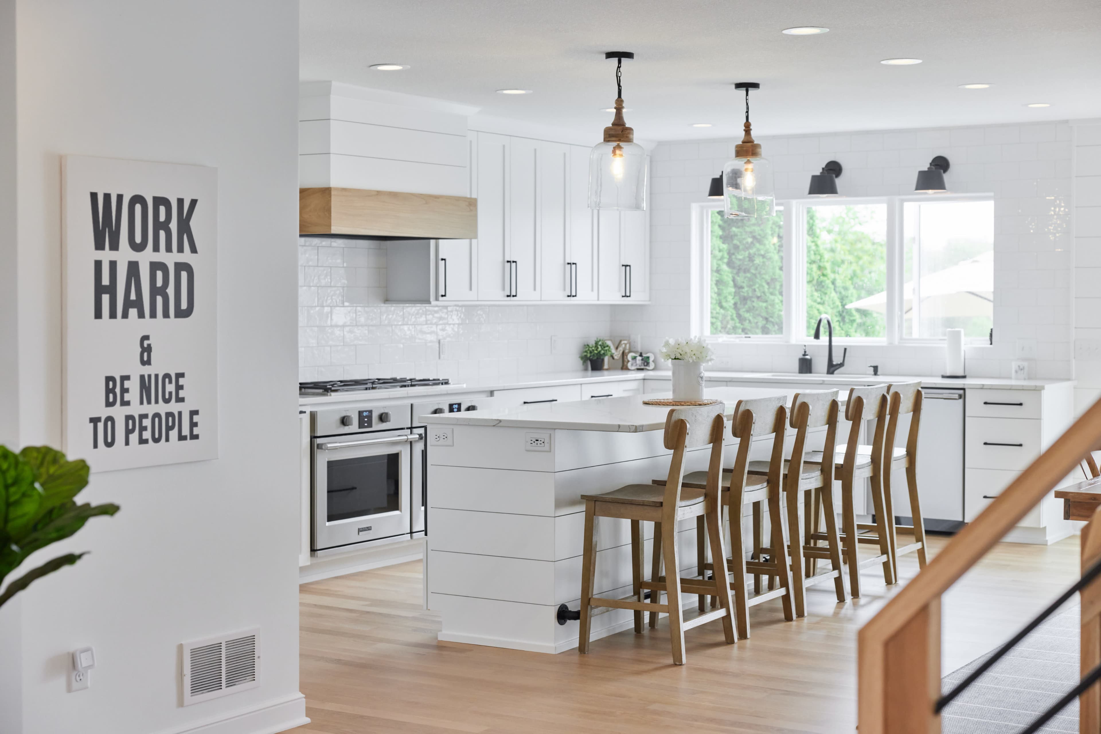 The image shows a modern kitchen featuring white cabinetry, a large island with wooden stools, and large windows letting in natural light.