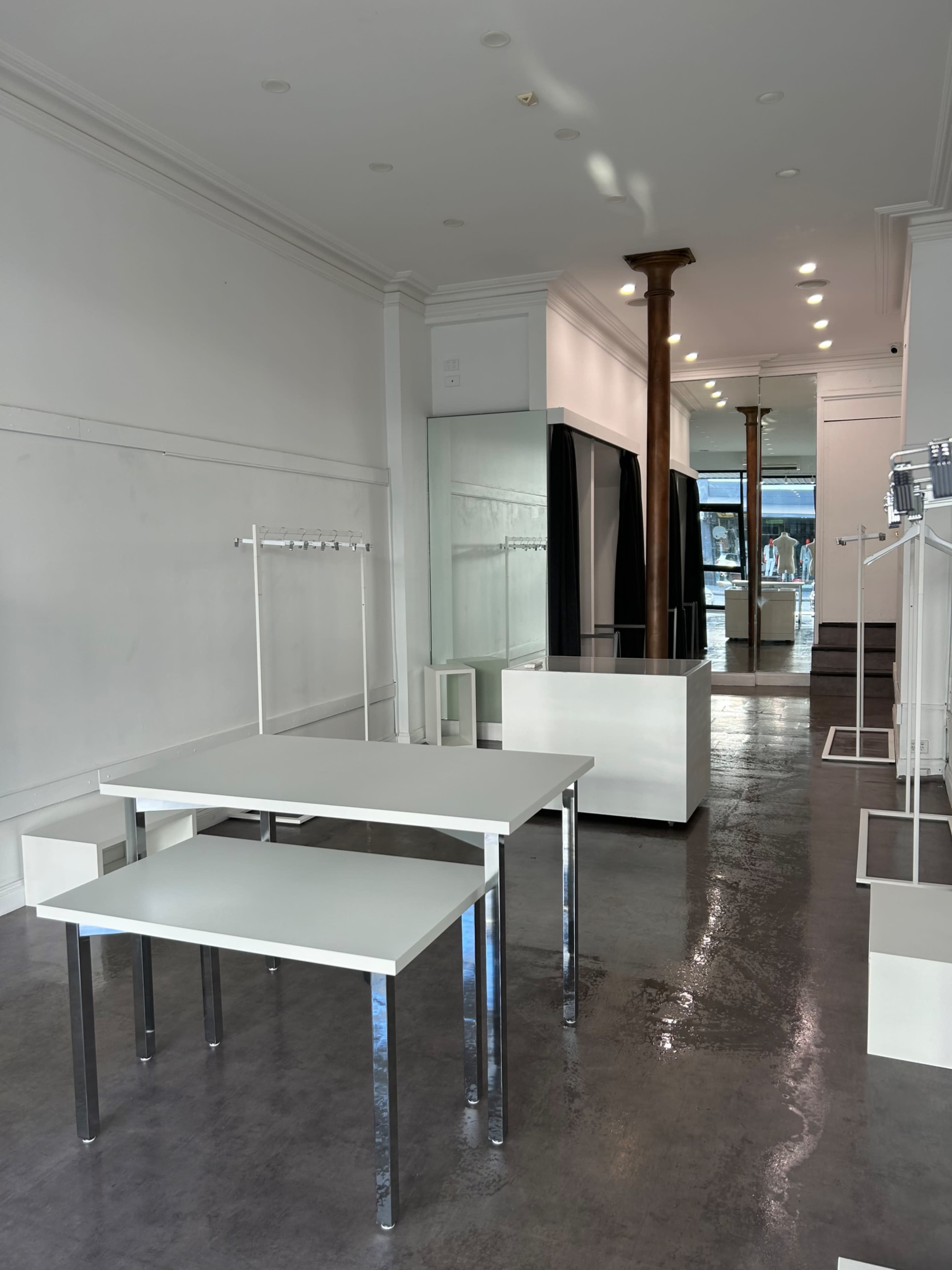 The image shows a minimalist retail space featuring white tables and display units with large windows letting in natural light.