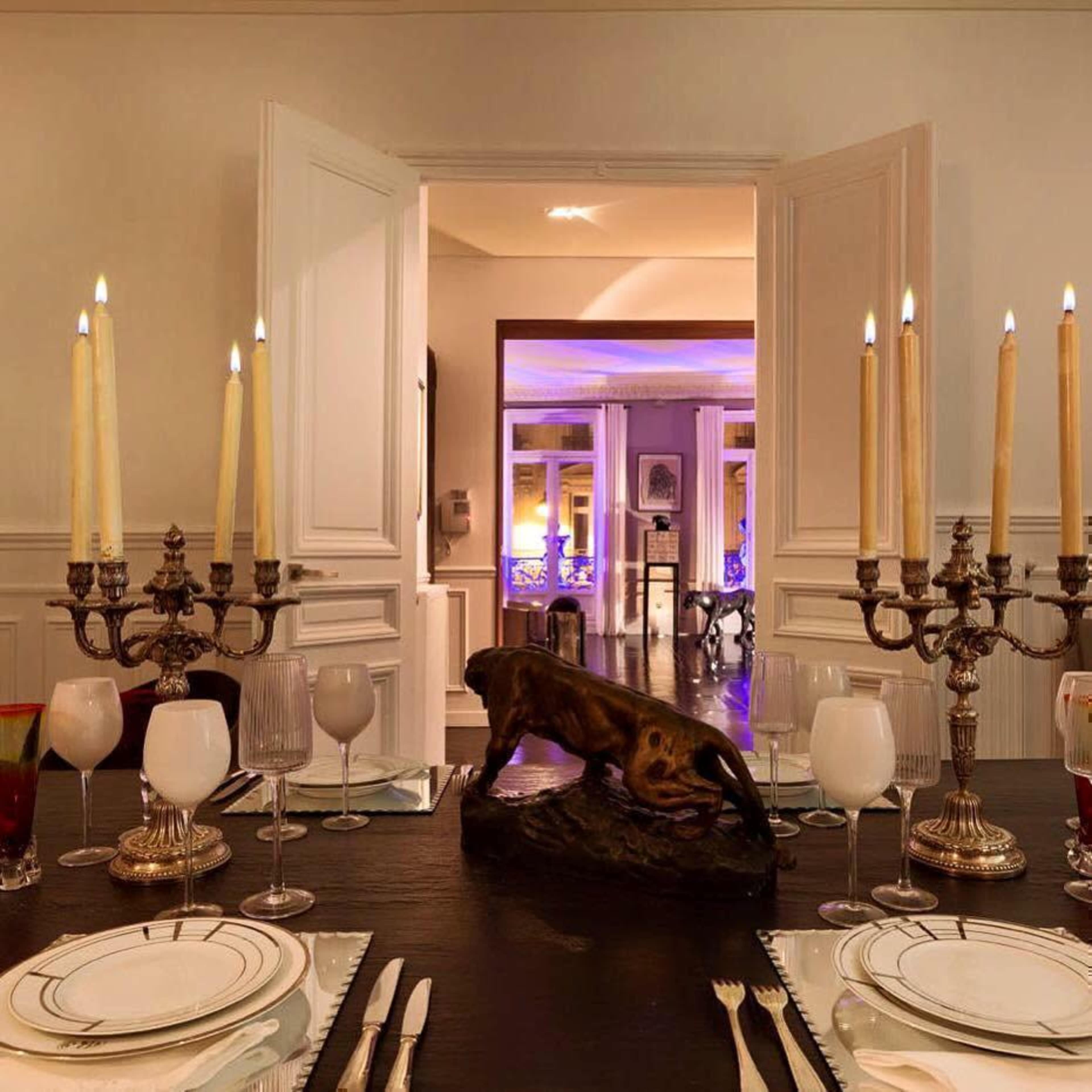 A well-decorated dining table set with candles, glassware, and cutlery, leading to a dimly lit room in the background.