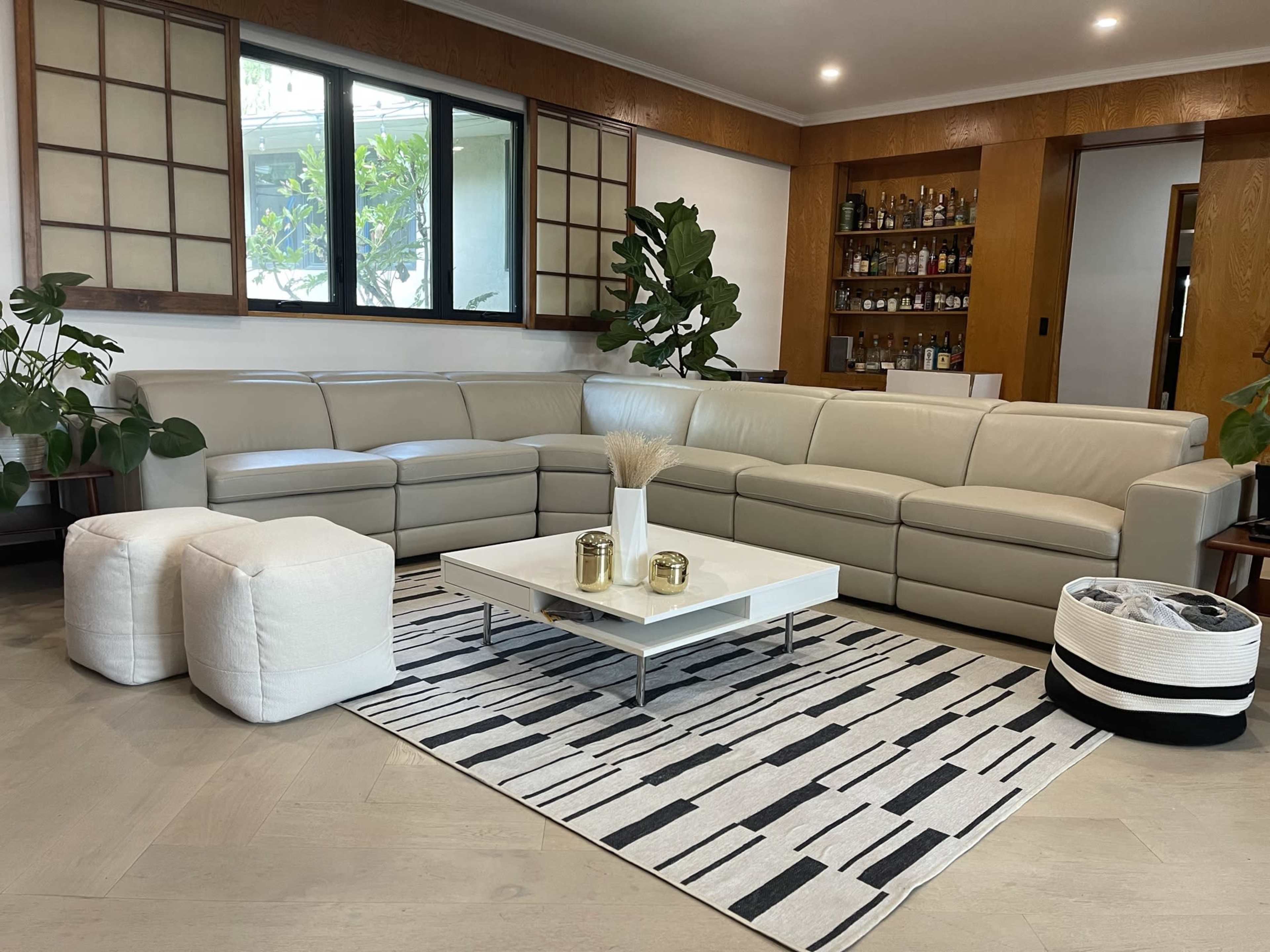 A modern living room featuring a light-colored sectional sofa, a white coffee table on a striped rug, and potted plants near large windows.