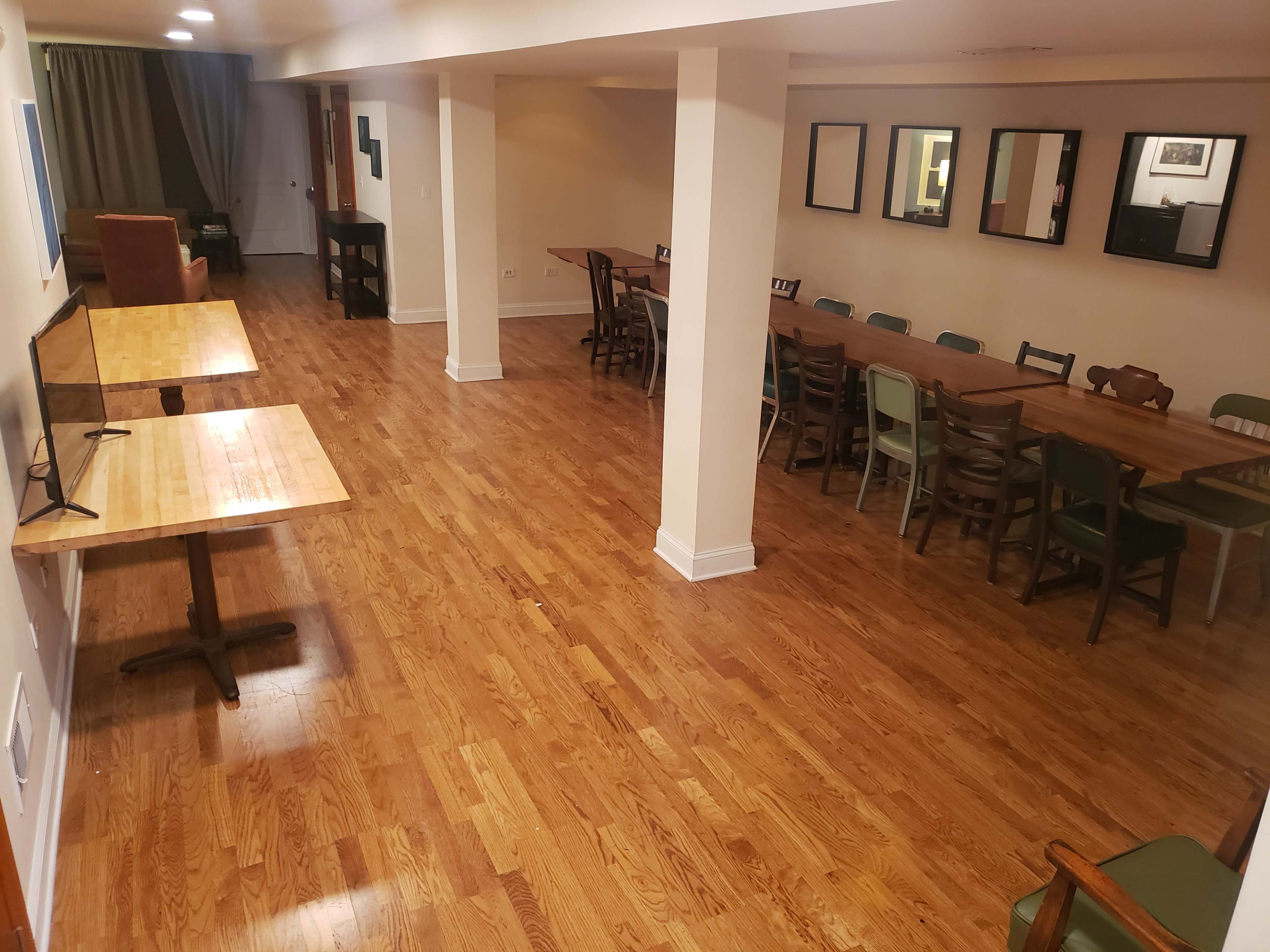 A spacious, well-lit room with wooden floors, featuring a long table, multiple chairs, and two smaller tables along one side.
